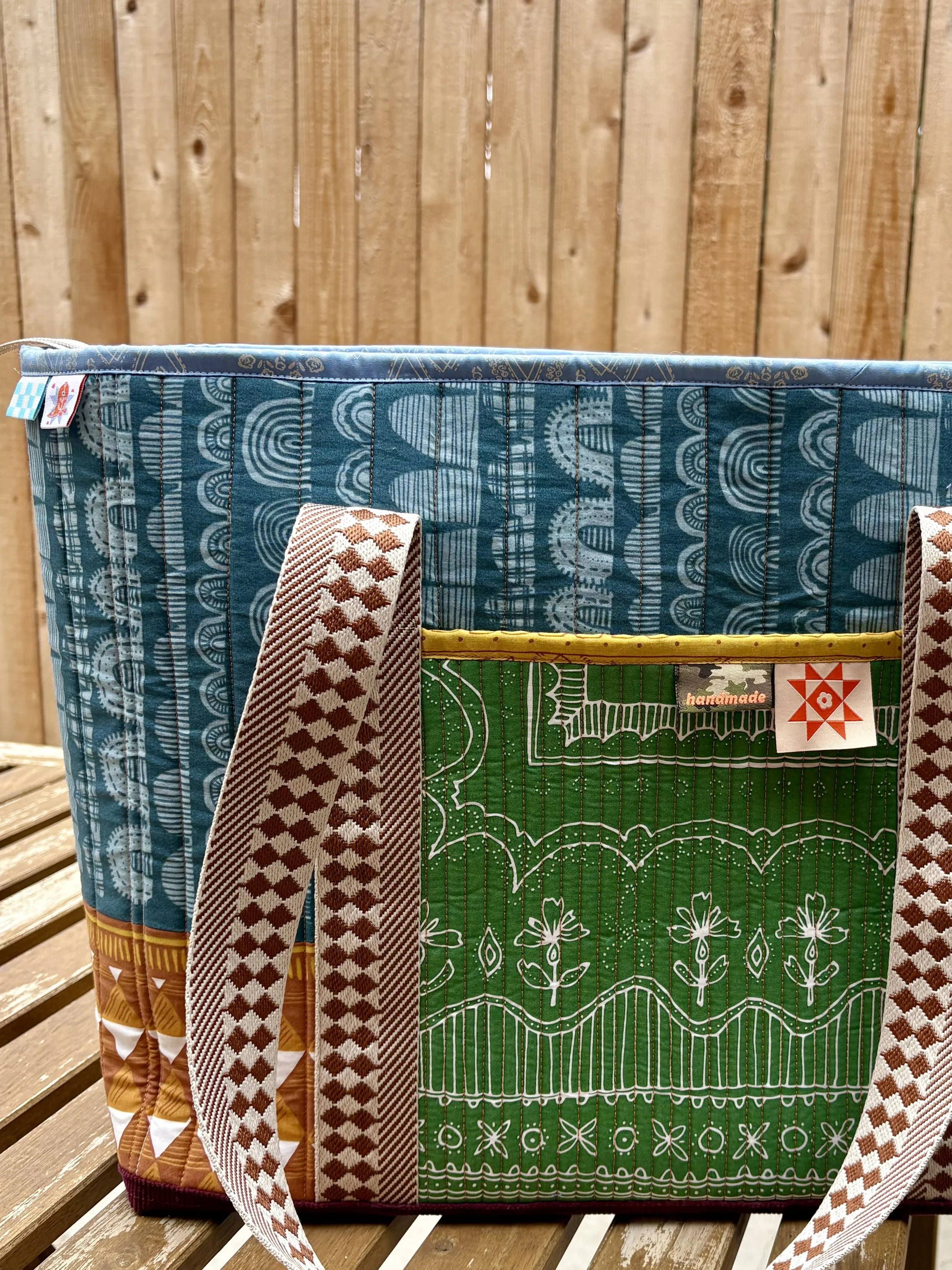 Handmade bohemian patchwork tote bag made from warm-toned bandana fabrics, with a green quilt block pocket and brown diamond-patterned straps.