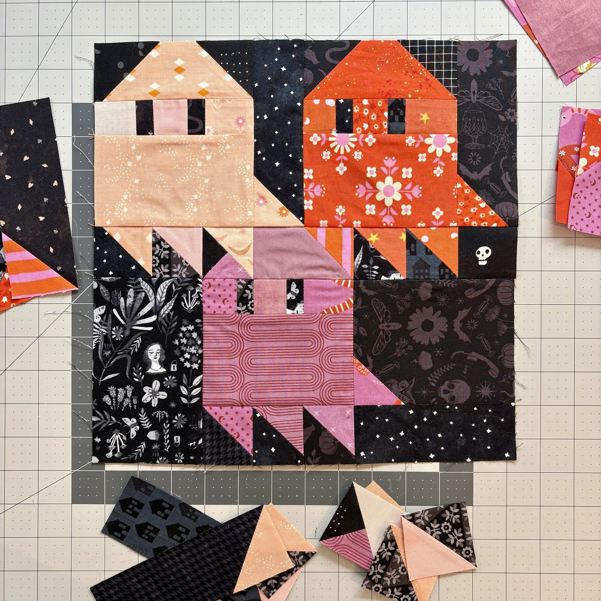 A quilt block with Halloween-themed fabric featuring two houses, a pumpkin, and a ghost, surrounded by black fabric with white patterns, with loose fabric pieces around it.