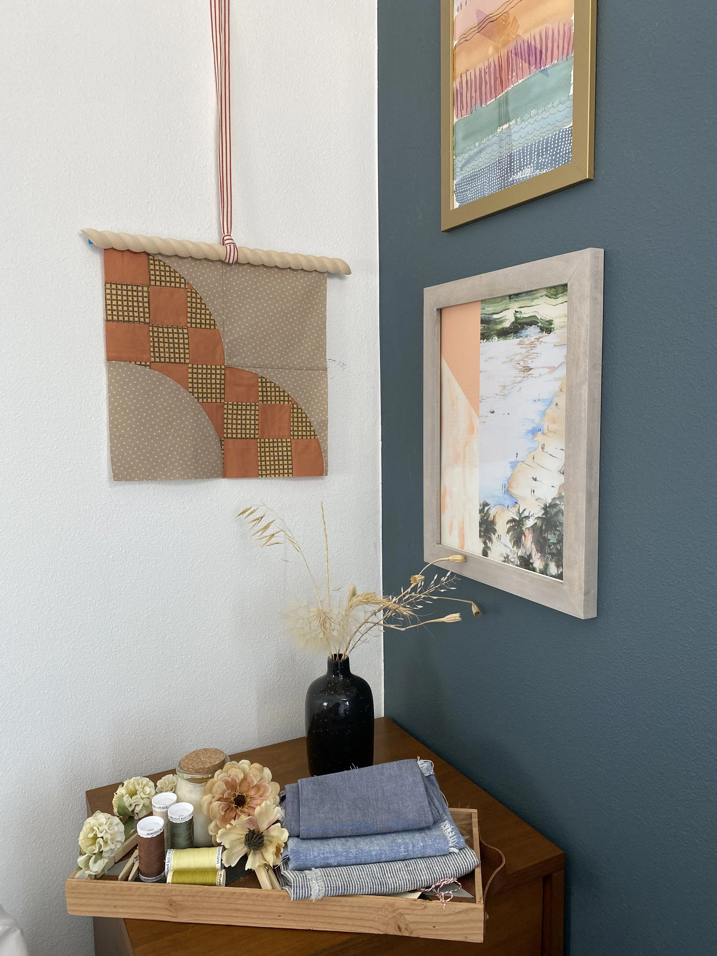 Decorative corner with framed artwork, a black vase with dried plants, a tray of sewing supplies including thread, fabric, flowers, and other crafting materials on a wooden table.