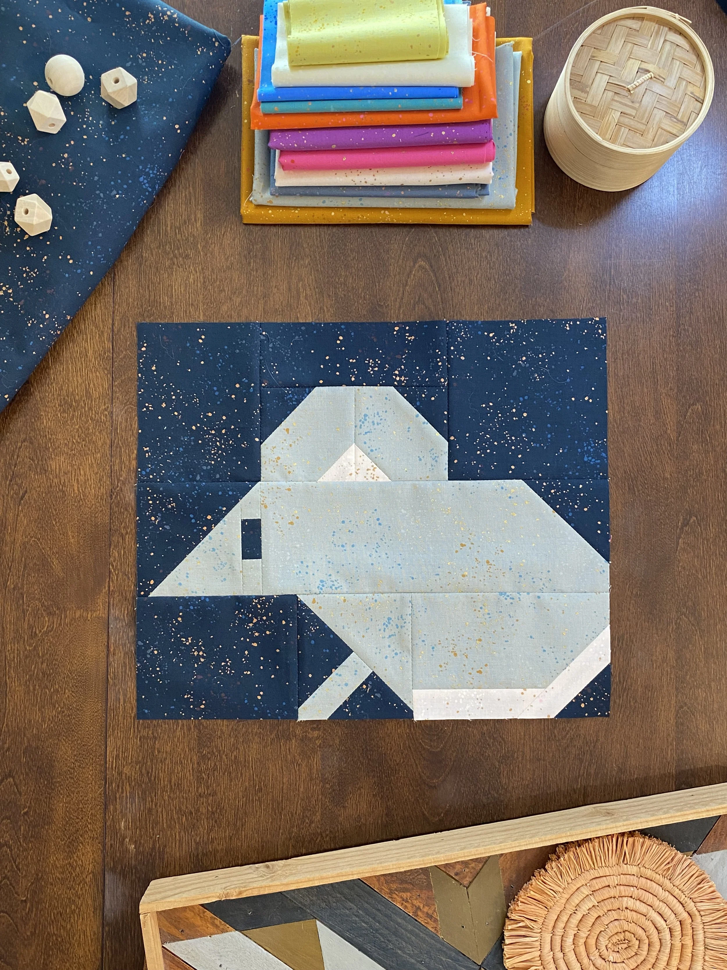 A quilt in progress with a geometric pattern of a white dog on a dark starry background, surrounded by sewing supplies and fabric on a wooden table.