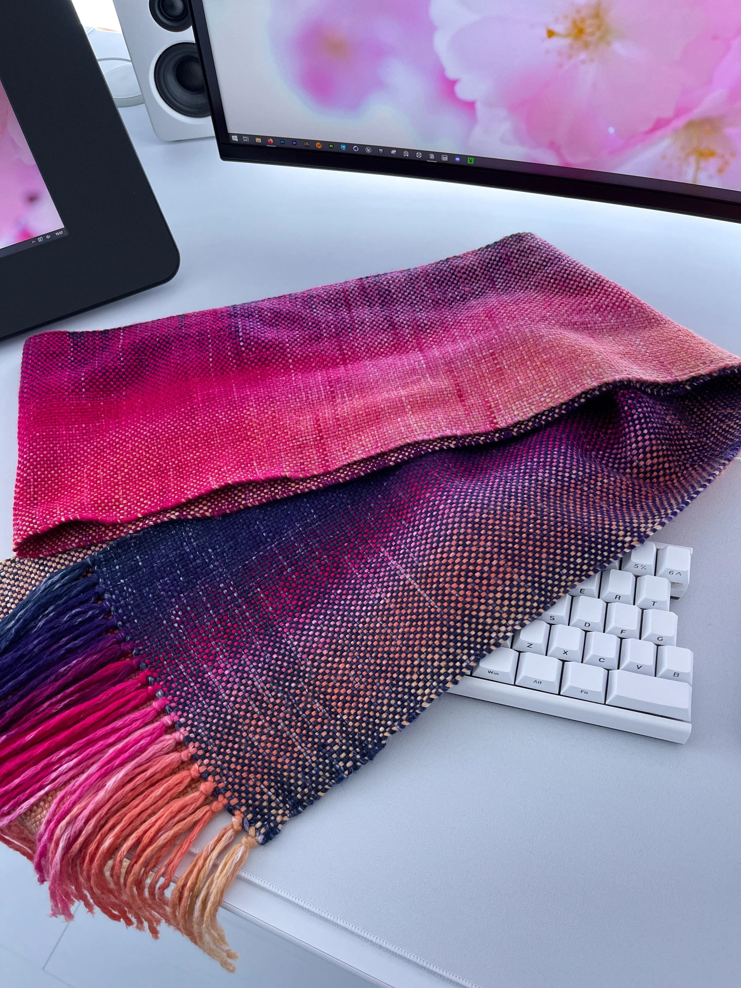 Hand-woven scarf of pink, purple, and orange hues resting on a white keyboard on white computer desk.