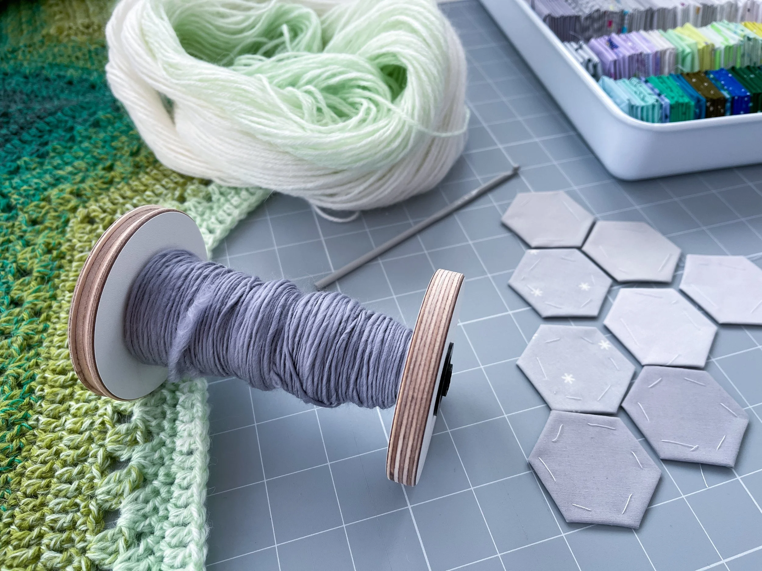 Bobbin of hand-spun grey fiber resting on top of large grid mat, with green crochet blanket, a crochet hook, and small, hand-stitched grey hexagons for quilting.