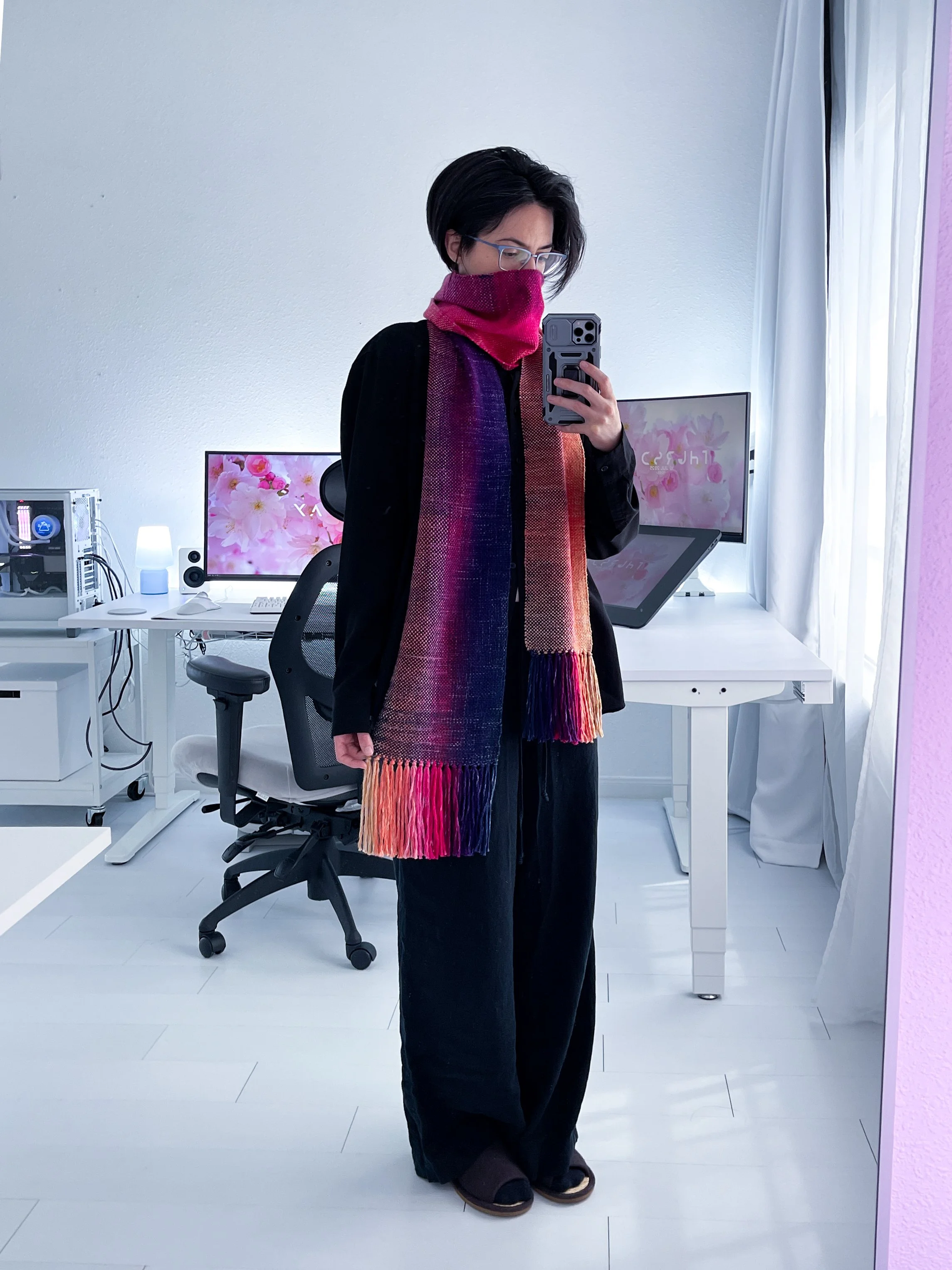 Hand-woven scarf of pink, purple, and orange hues draped over figure in black standing in an office with white floors and walls, with computer desk in the background.