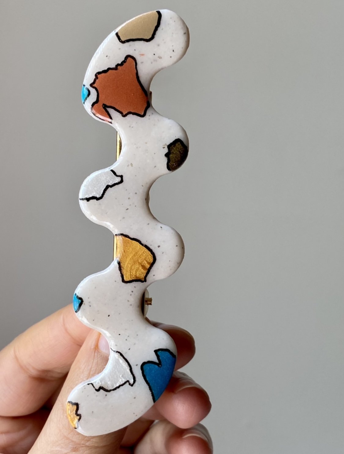 Polymer clay Hand holding a wavy, abstract brooch with multicolored patches on a white background.