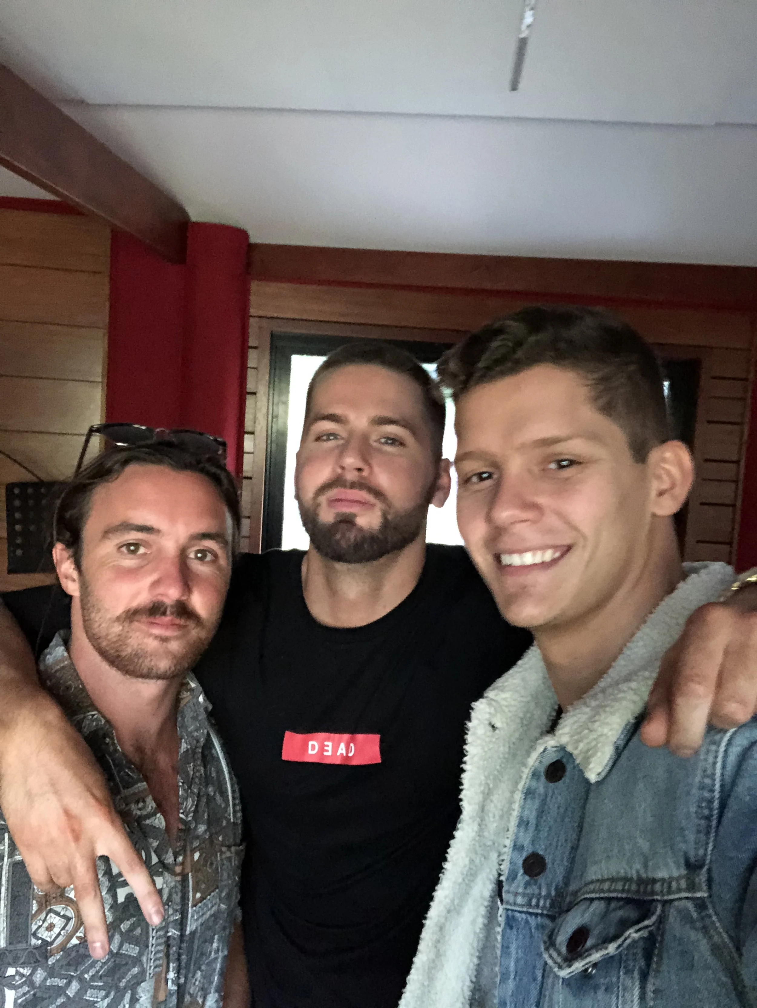 Three men smiling and posing for a photo indoors, with wooden walls and red curtains in the background.