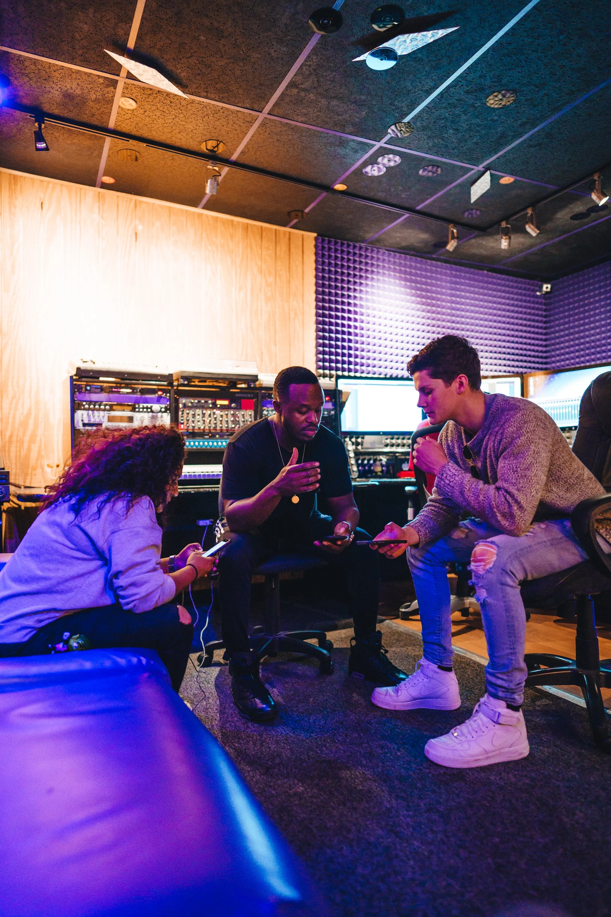 Three people in a music studio with sound equipment and computer monitors, engaged in conversation or collaboration.