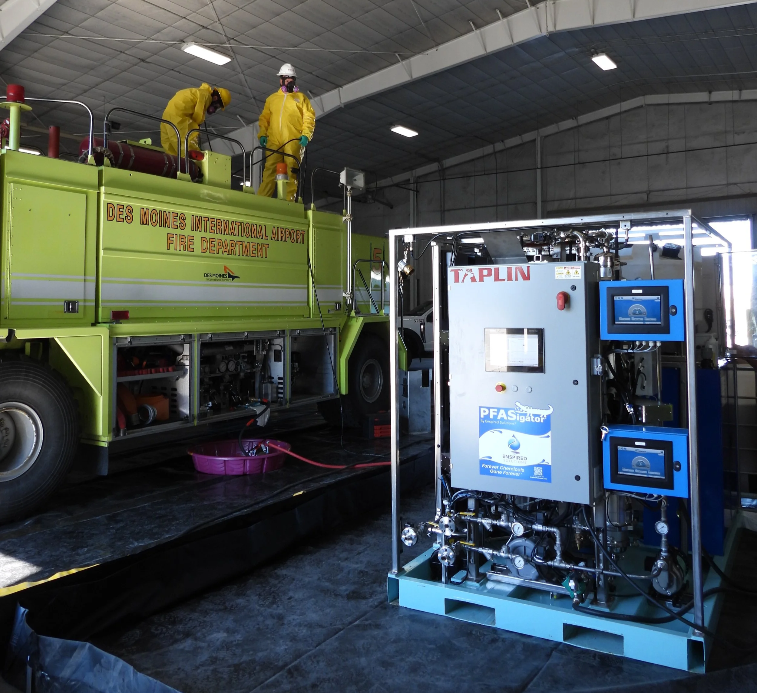 Firefighters in yellow hazmat suits working on a fire truck labeled 'Des Moines International Airport Fire Department' inside a warehouse, with equipment and monitors nearby.