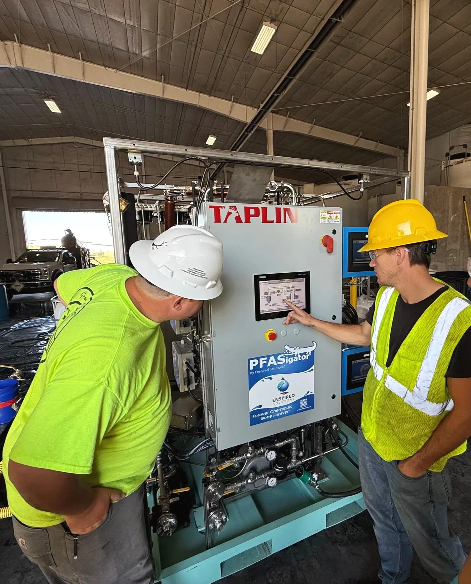Two workers in safety helmets and vests examining and operating a control panel on a large industrial machine inside a warehouse or factory.
