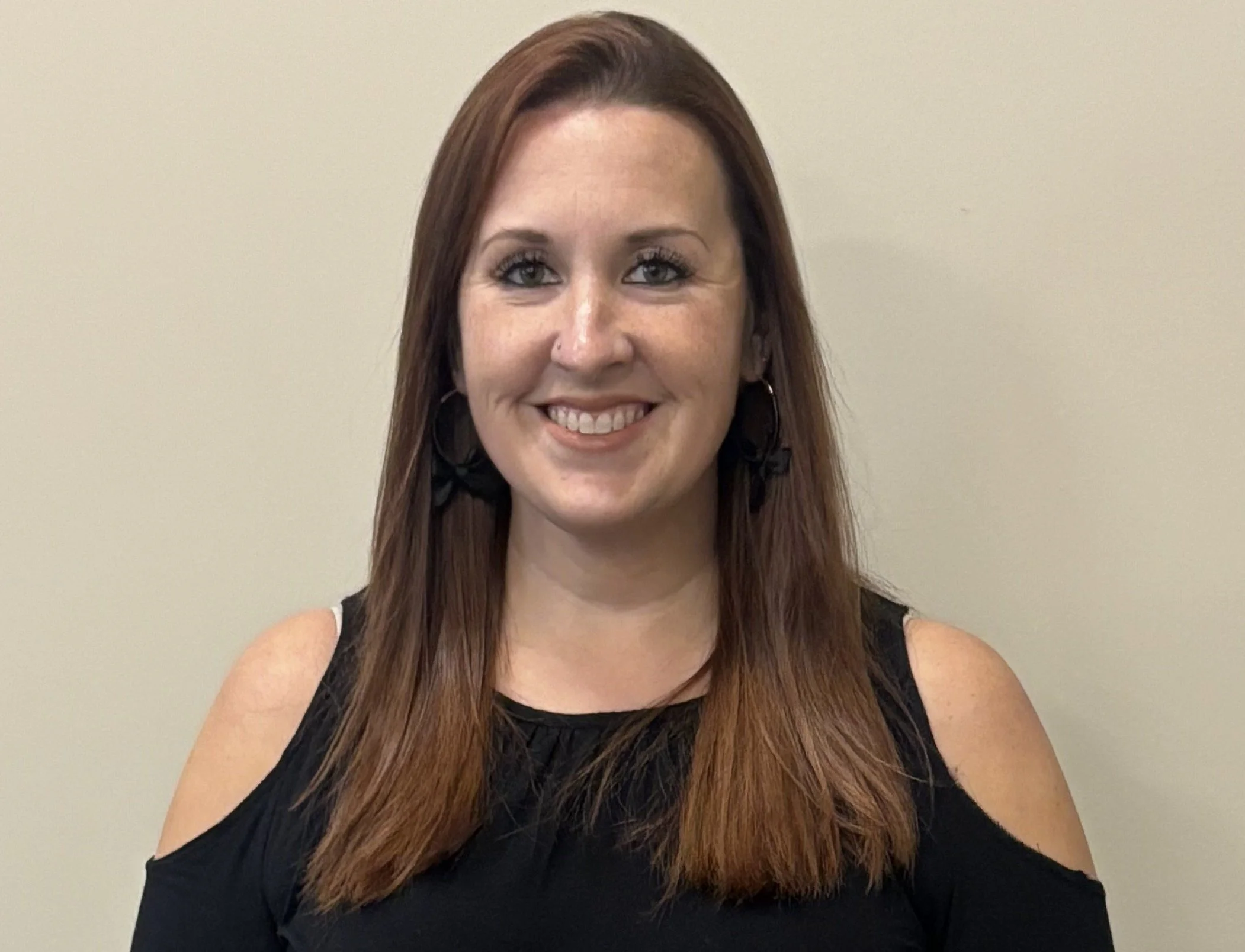 A woman with long reddish-brown hair smiling at the camera, wearing a black cold-shoulder top and black earrings, standing against a plain beige wall.