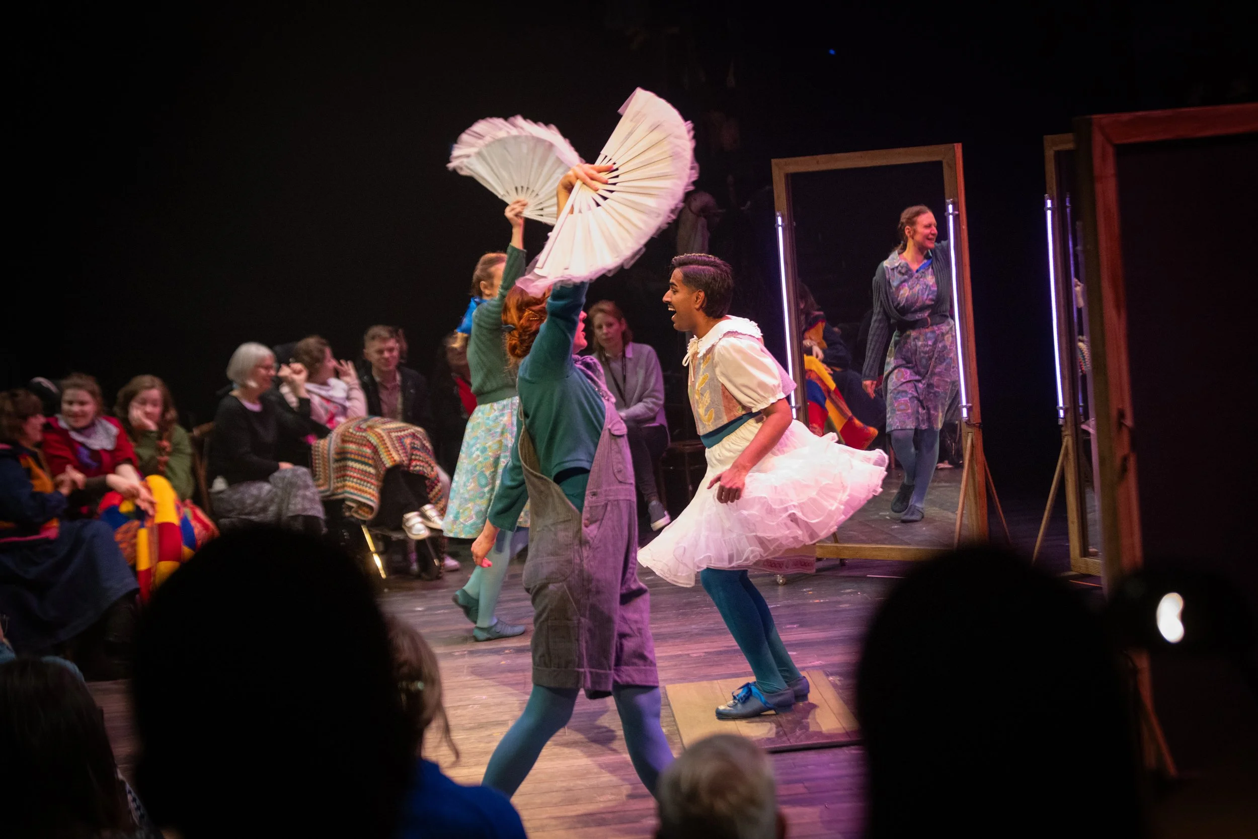 People performing a dance or theatrical scene on stage, with an audience watching. The performers are dressed in colorful costumes, some holding fans, and are engaged in expressive movements and interactions.