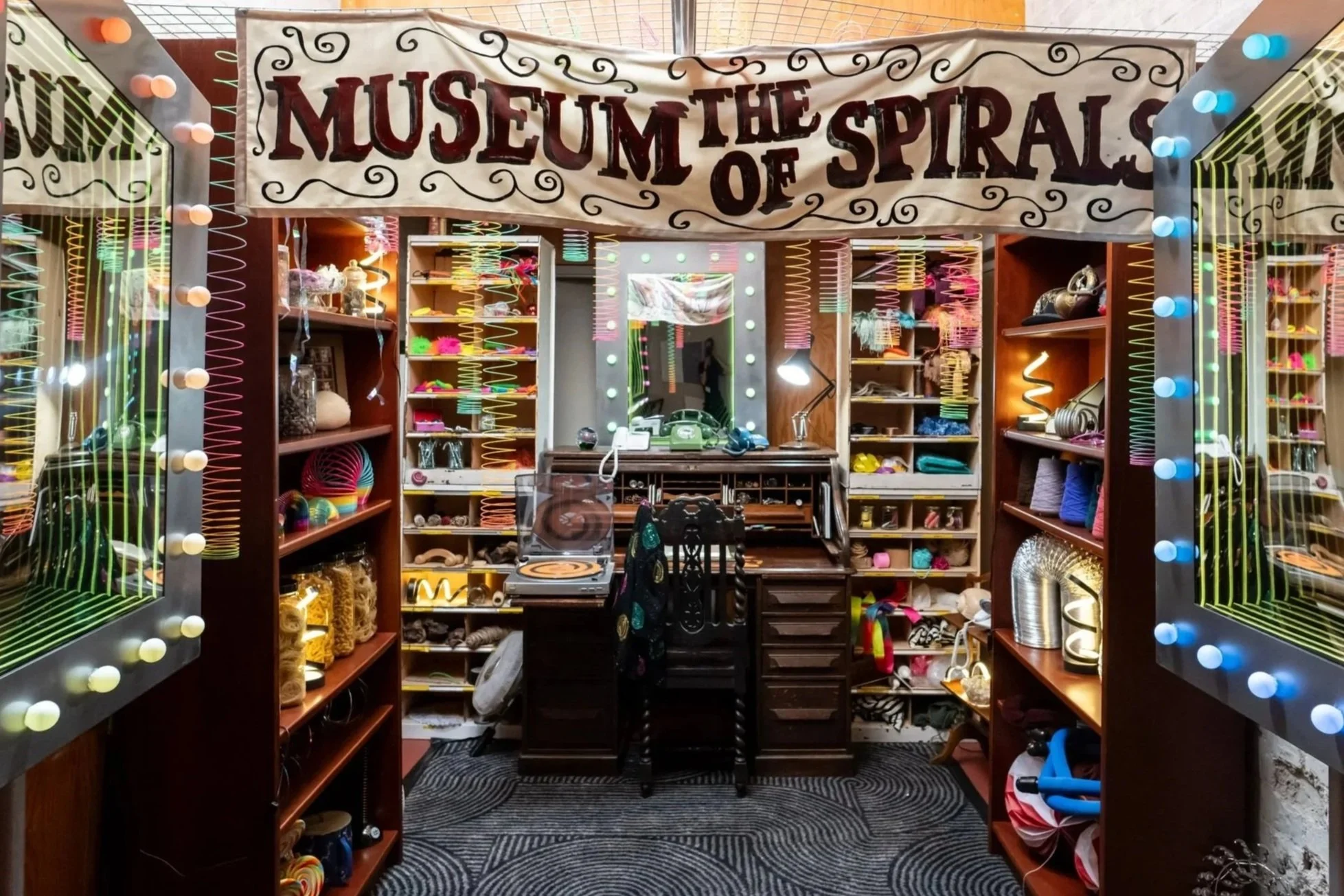Interior of a bedroom or art studio with shelves filled with color and craft supplies, a large mirror framed with lights, a vintage green rotary phone on a wooden desk, and a spinning turntable in the foreground.
