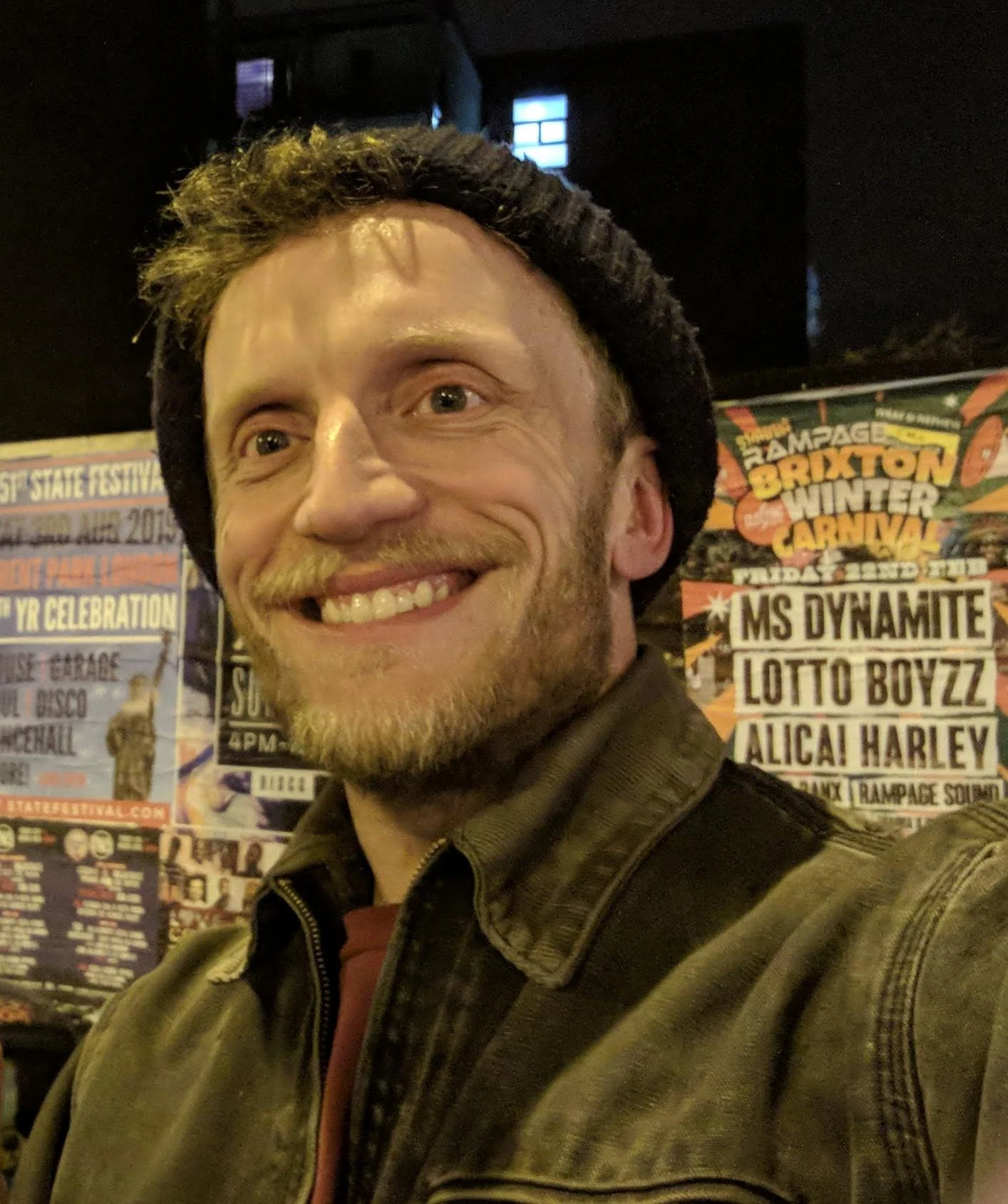 A smiling man with a beard, wearing a black beanie and a brown jacket, taking a selfie at a festival or concert with a colorful poster behind him listing performances and events.