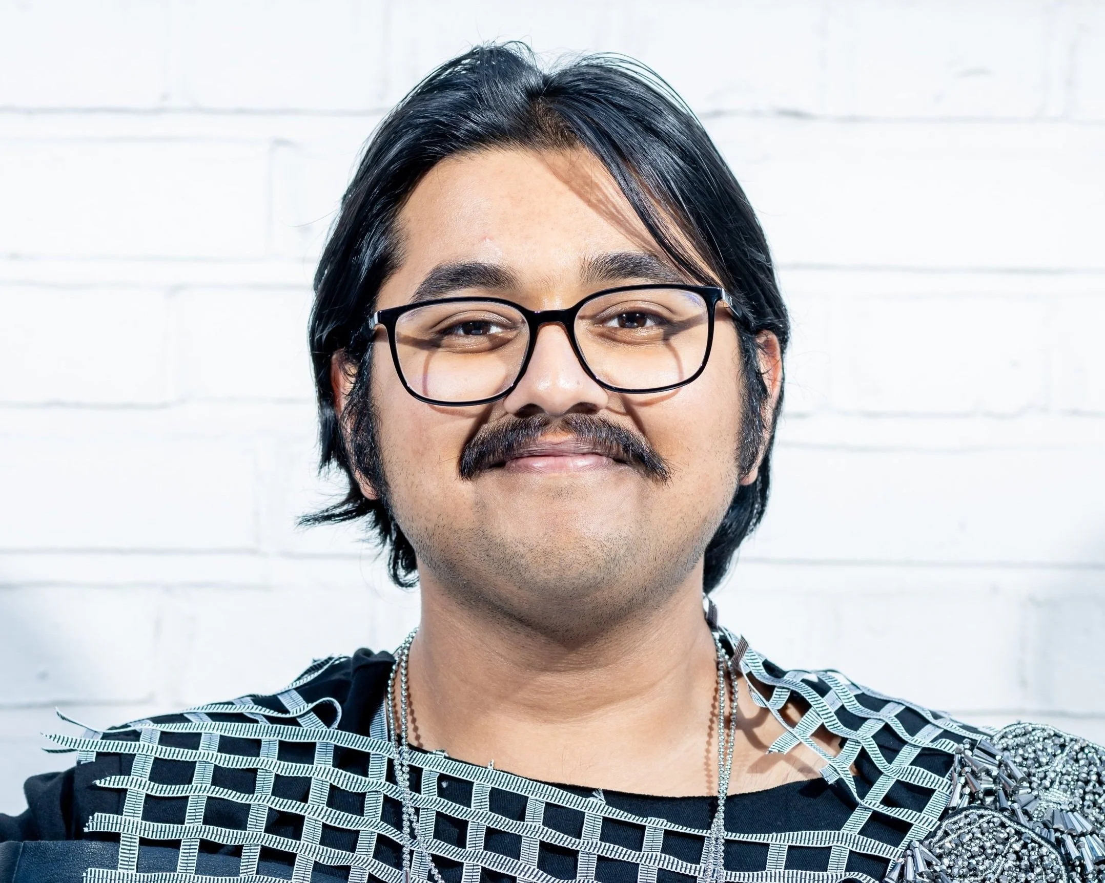 A man with black hair, glasses, and a mustache, smiling, standing in front of a white brick wall.