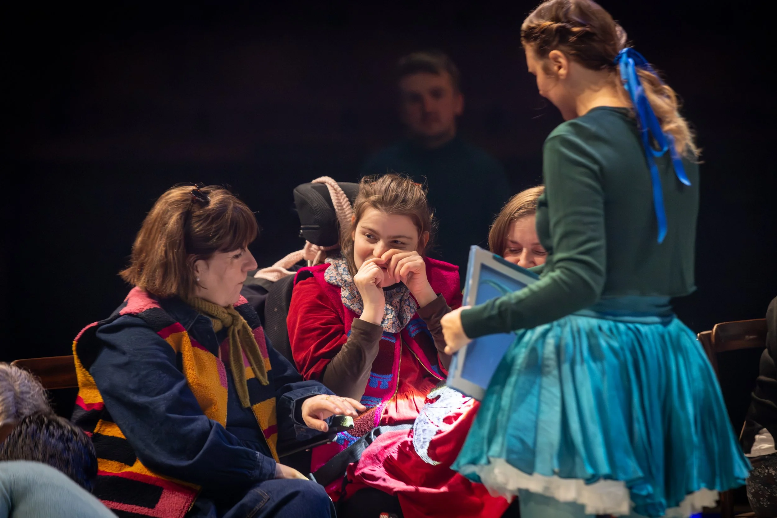 A group of women sitting in a theater or lecture hall, engaged in conversation while a young girl standing in front of them appears to be showing or explaining something to the group.