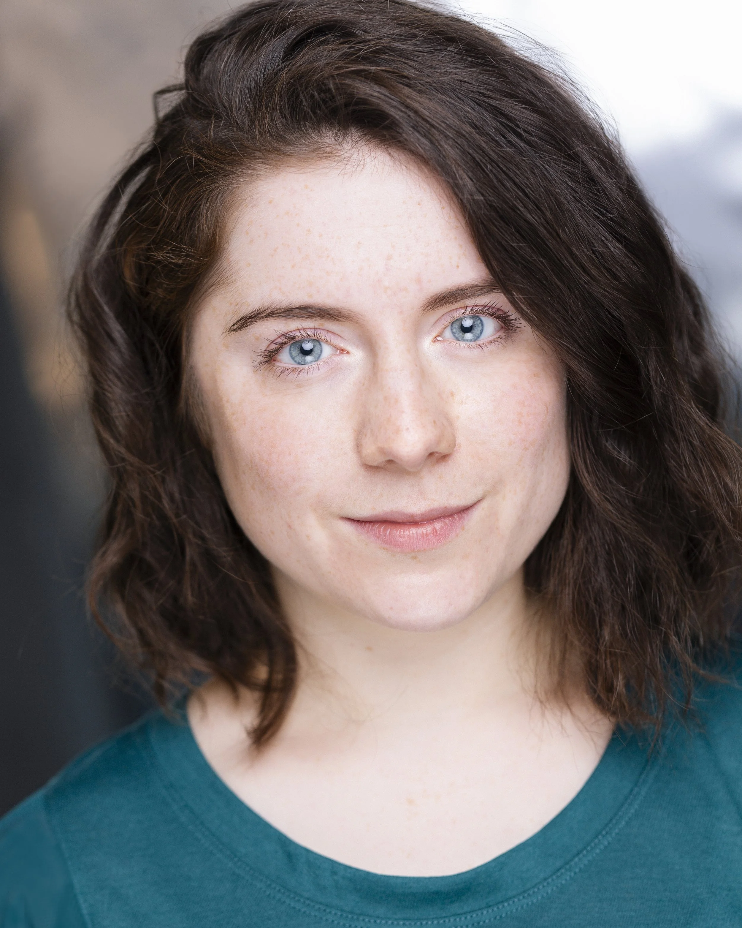 Close-up portrait of a young woman with shoulder-length wavy brown hair and blue eyes, wearing a teal shirt.