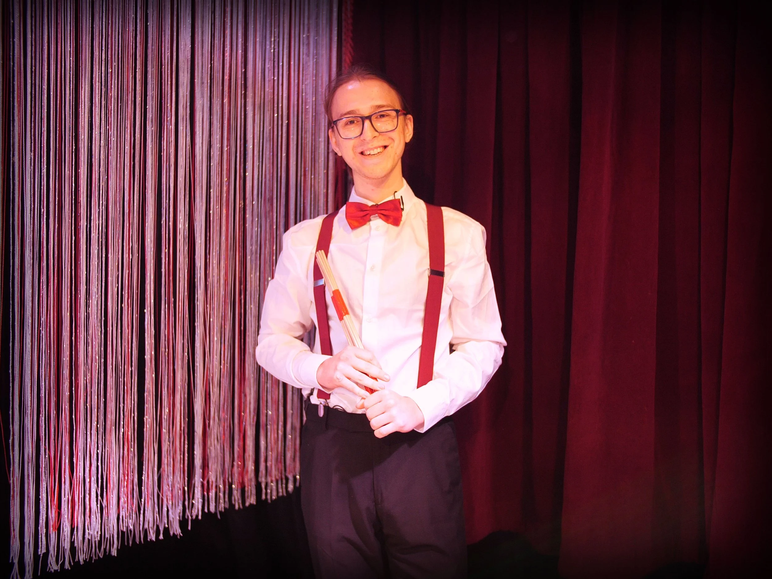A smiling man dressed as a magician, wearing glasses, a white shirt, red bow tie, suspenders, and holding a magic wand, standing in front of a stage with red curtains and decorative fringe.