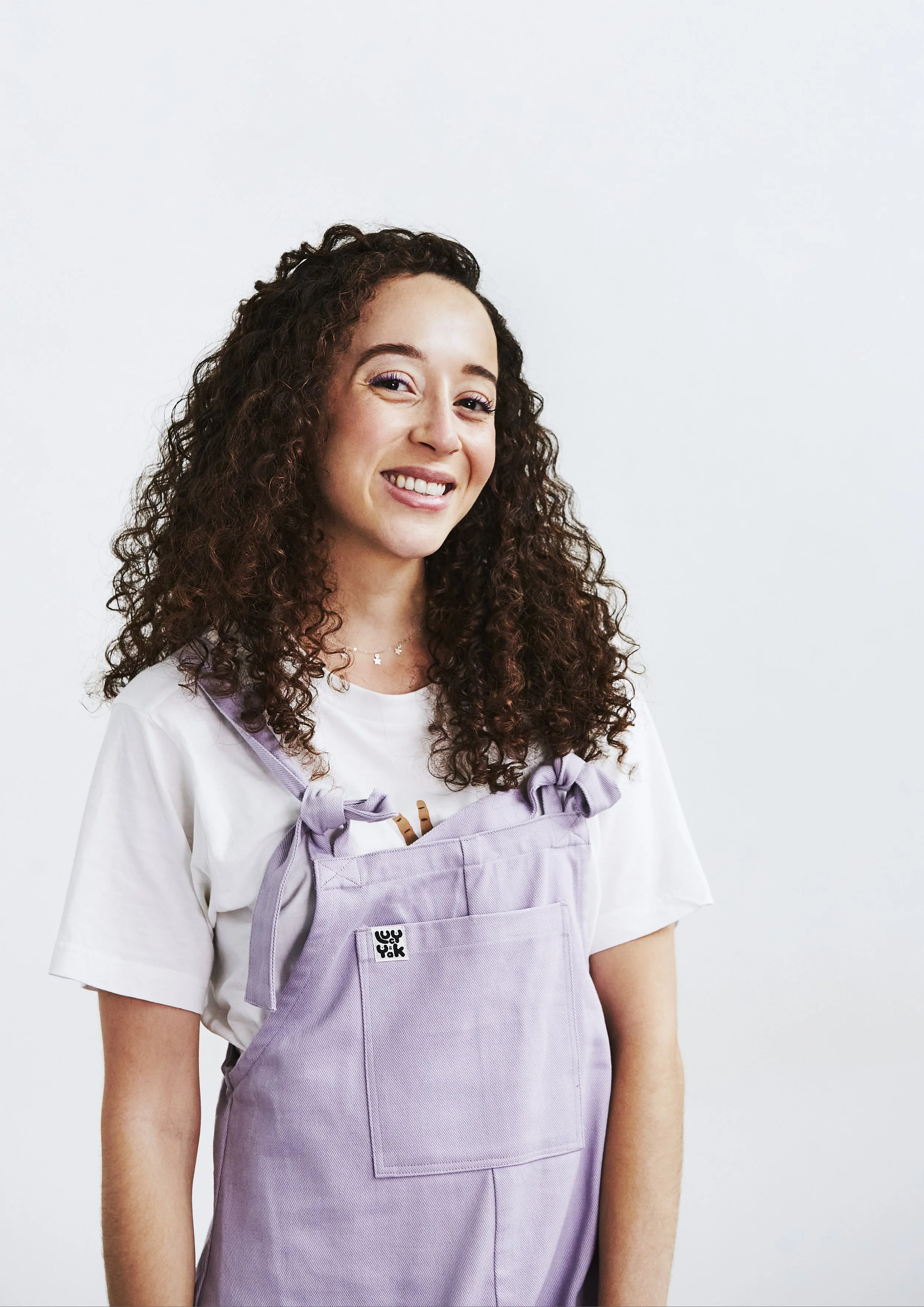 Smiling young woman with curly hair wearing a white shirt and a lavender apron, standing against a white background.