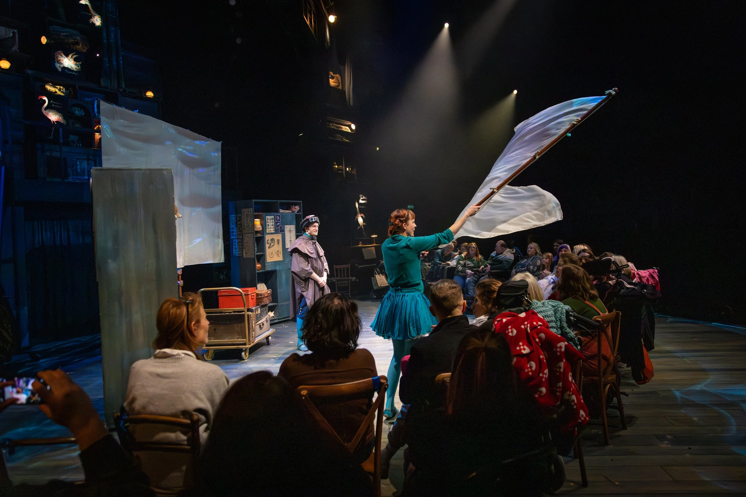 Stage performance with actors in costume, theater audience seated watching, stage props including a canvas and shelves, atmospheric lighting with spotlights.