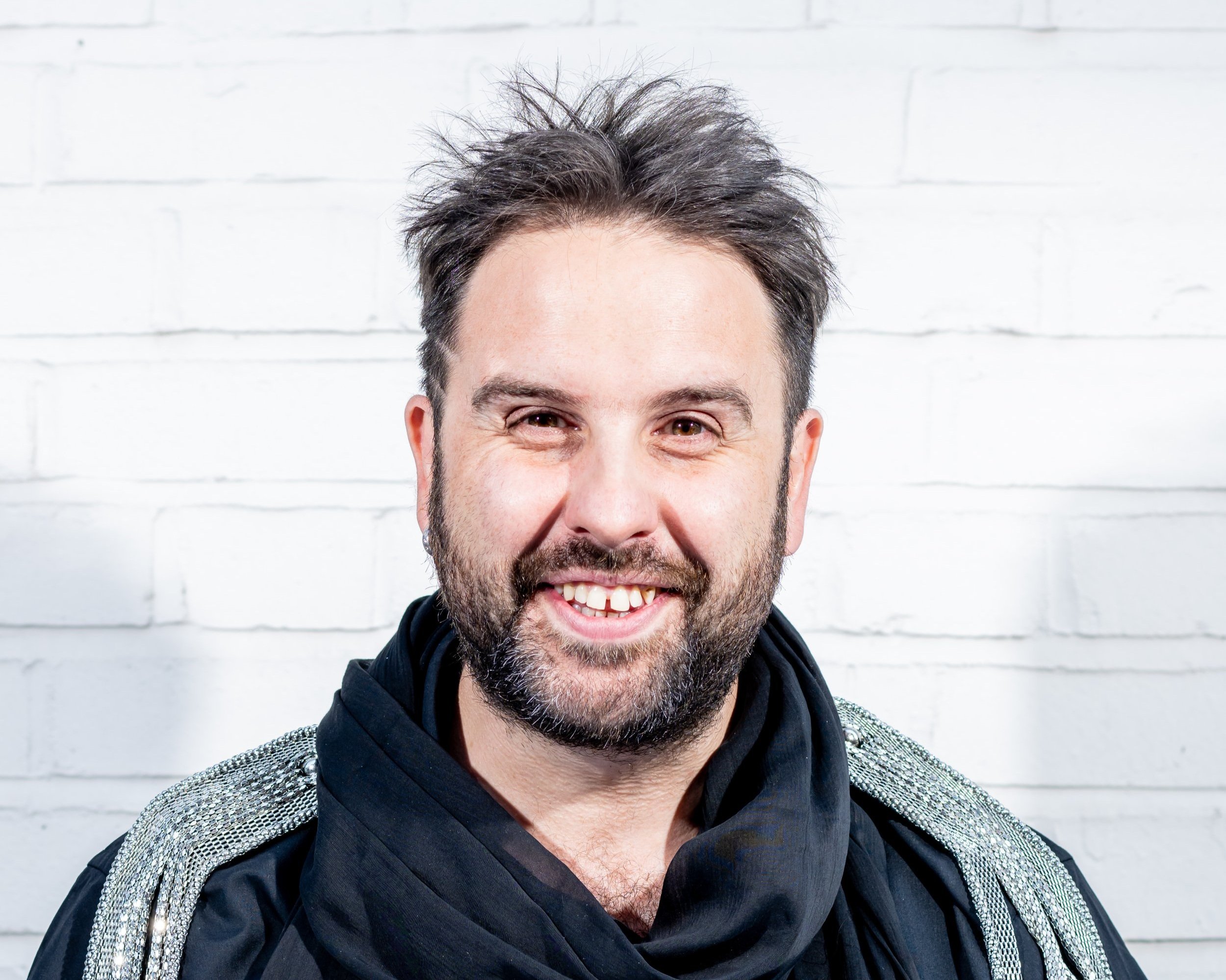 A smiling man with dark hair, a beard, and a mustache, wearing a black scarf and a jacket with metallic embellishments, standing against a white brick wall.