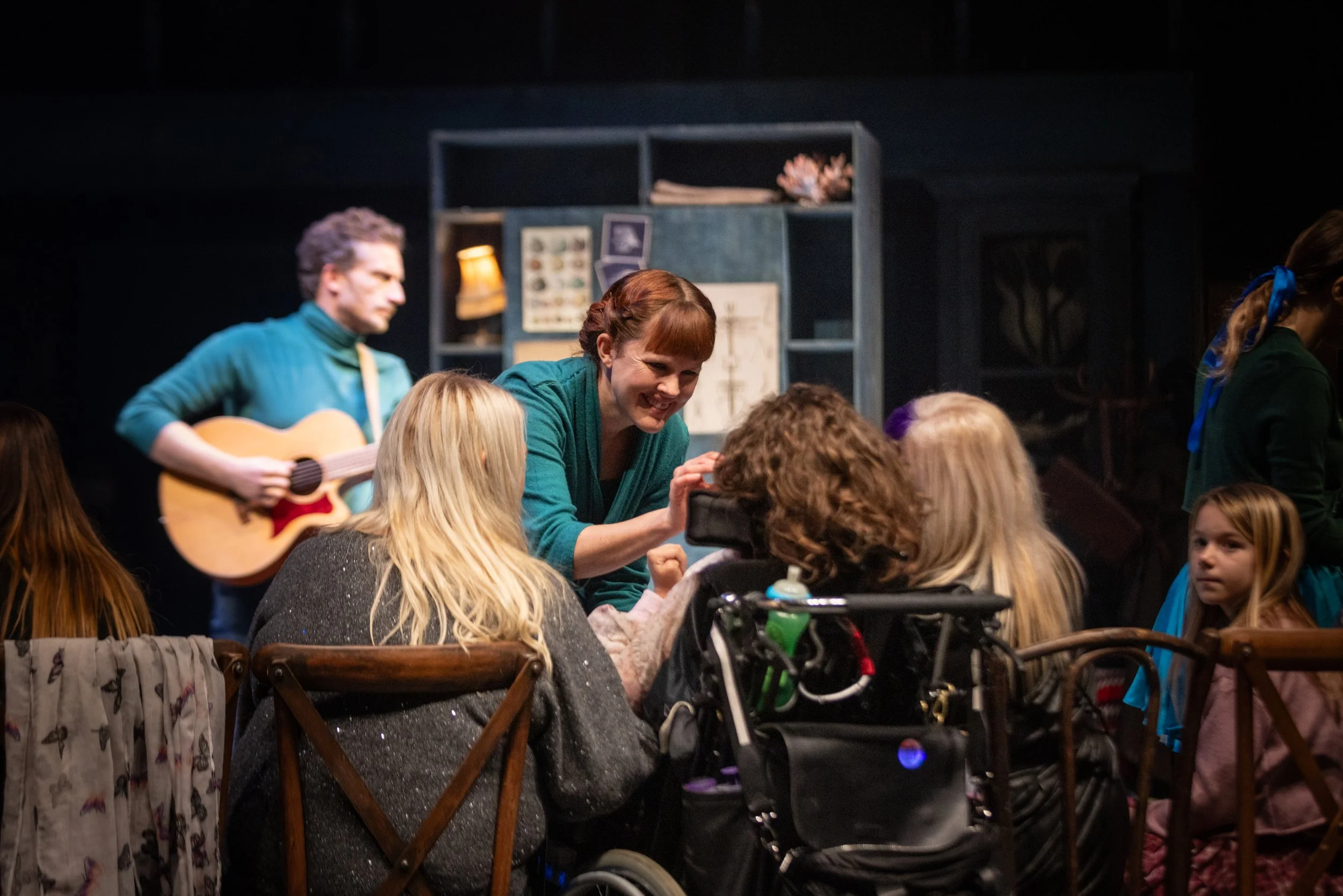 People gathered in a room, some sitting and some standing, with one person playing guitar. The atmosphere appears lively and engaging.