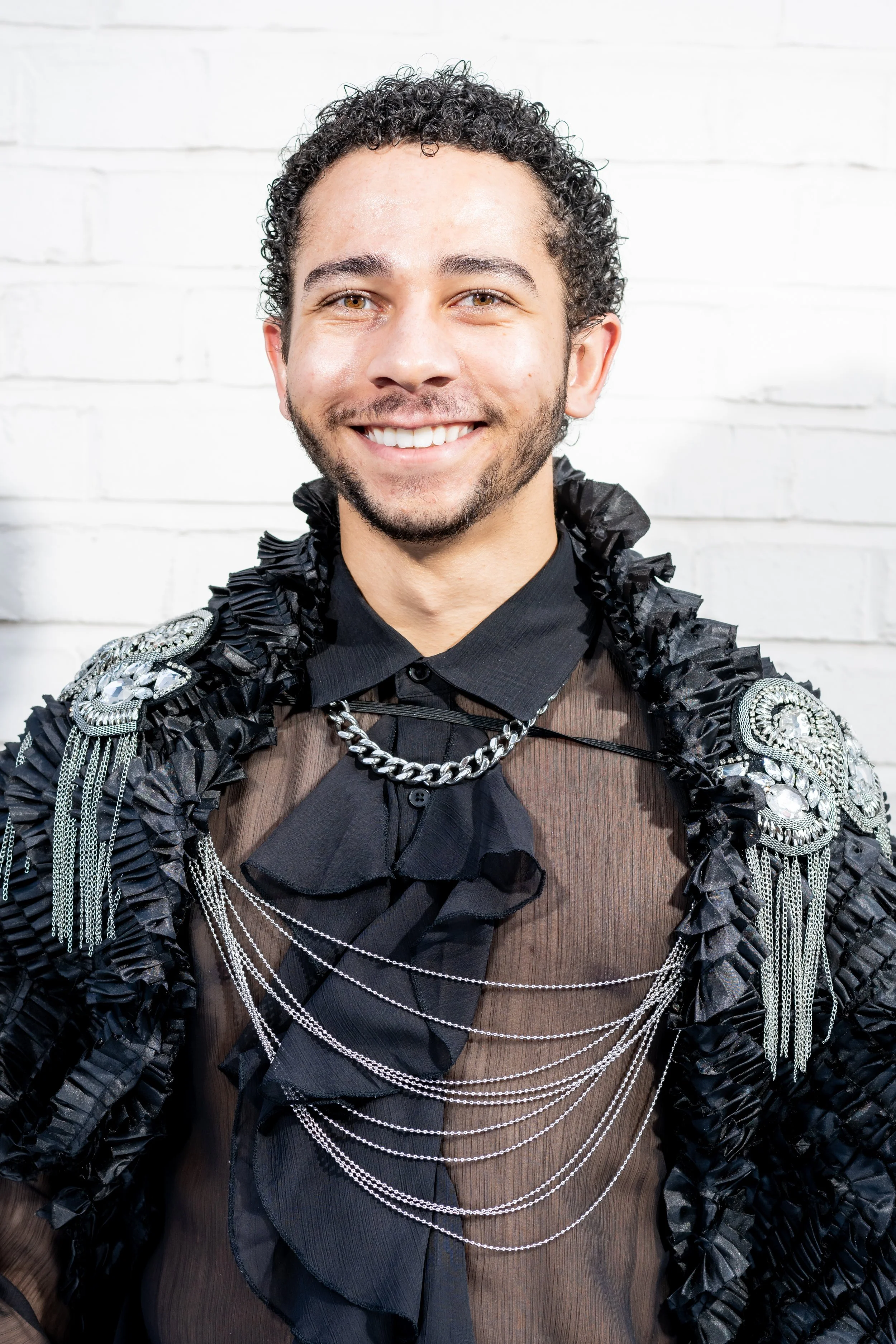 A smiling man with curly hair and a beard wearing a sheer black shirt with ruffles, silver chains, and decorative embellishments on his jacket, standing in front of a white brick wall.
