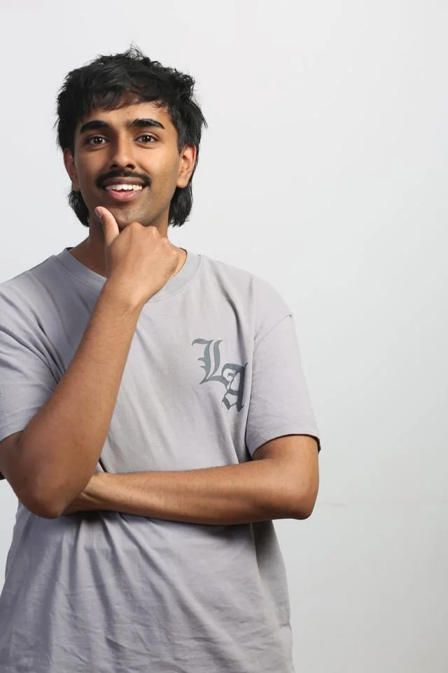 A young man with dark hair and a mustache, smiling, wearing a light gray T-shirt, standing against a plain white background.