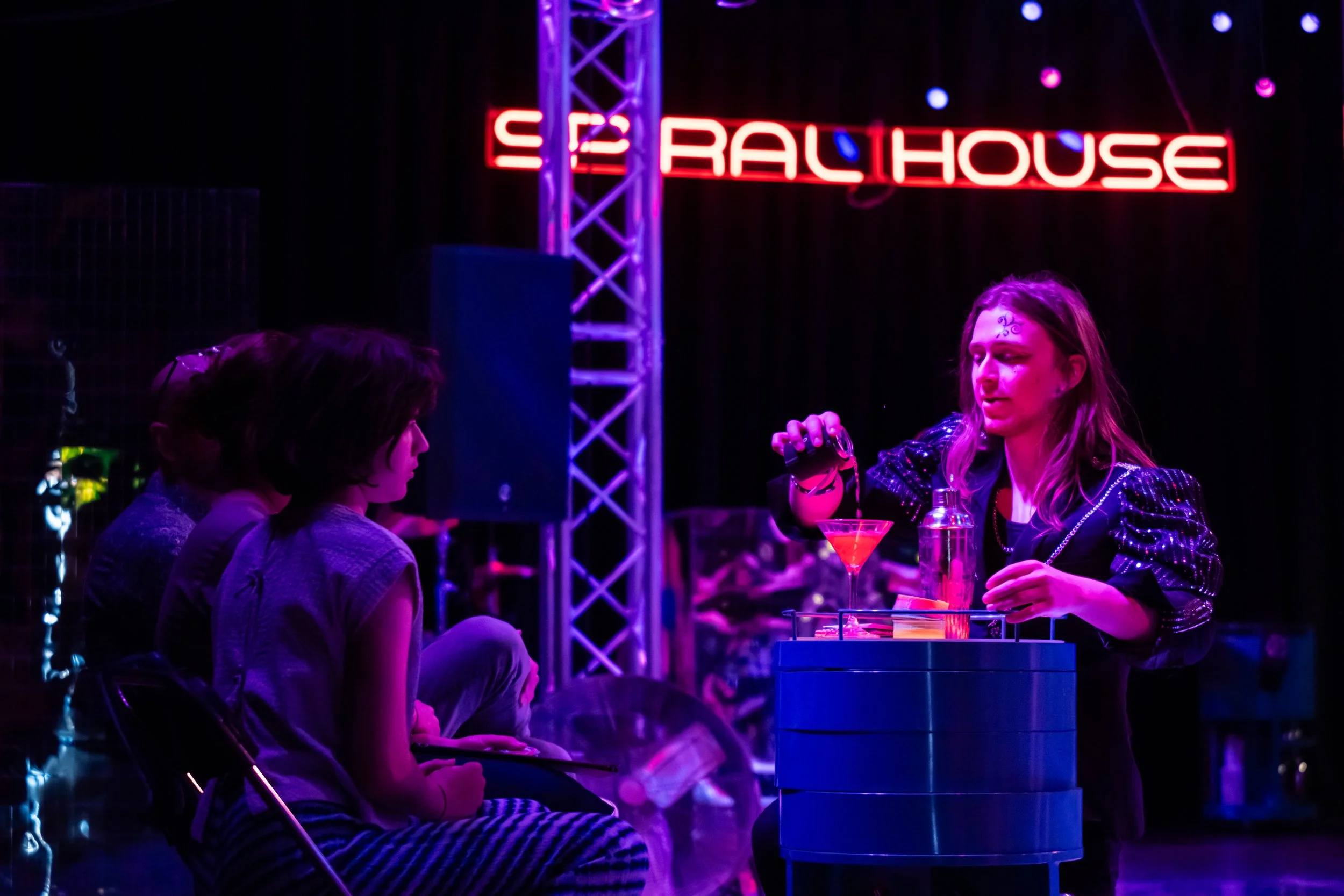 Two women sitting at a table, having a conversation in a dark, neon-lit nightclub or bar. There is a neon sign that reads 'ELECTRA CLUB RAL HOUSE' in the background, with purple and red lighting and hanging decorative lights.