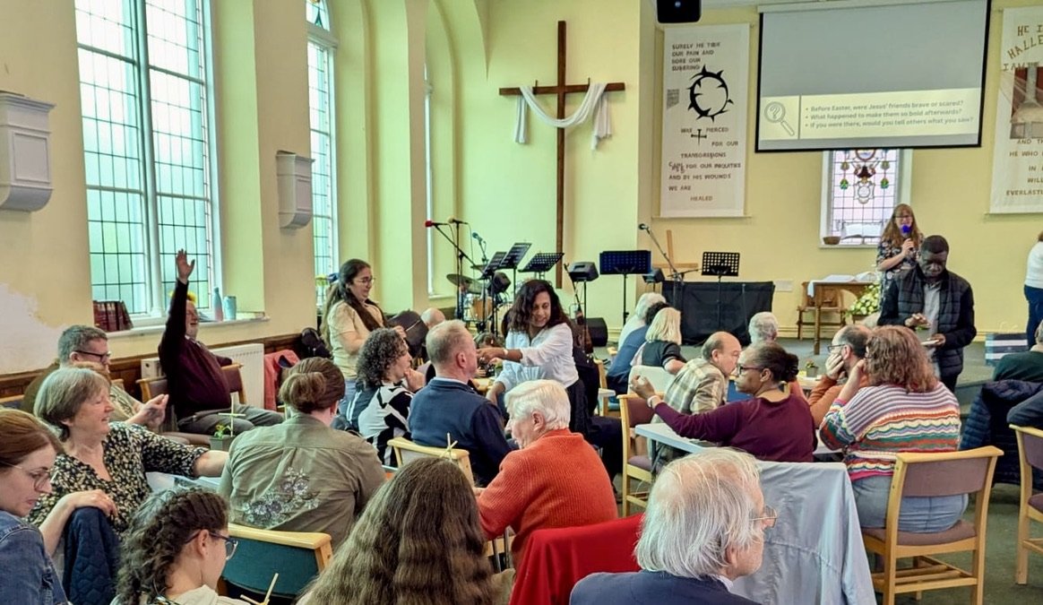 People gathered inside a church during a meeting or event, with some raising their hands, seated around tables, and a woman standing near the front speaking into a microphone. There are musical instruments and microphones on stage, with a large cross