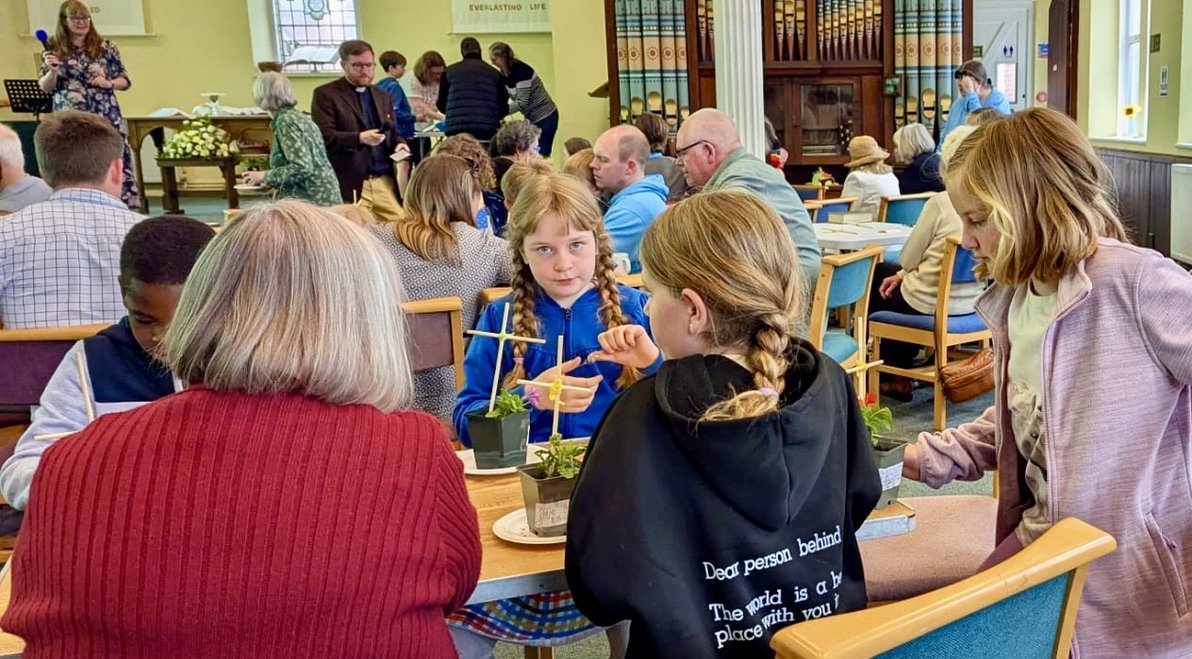 People sitting at tables in a church or community hall, engaging in conversation and activities, with some planting small potted plants.