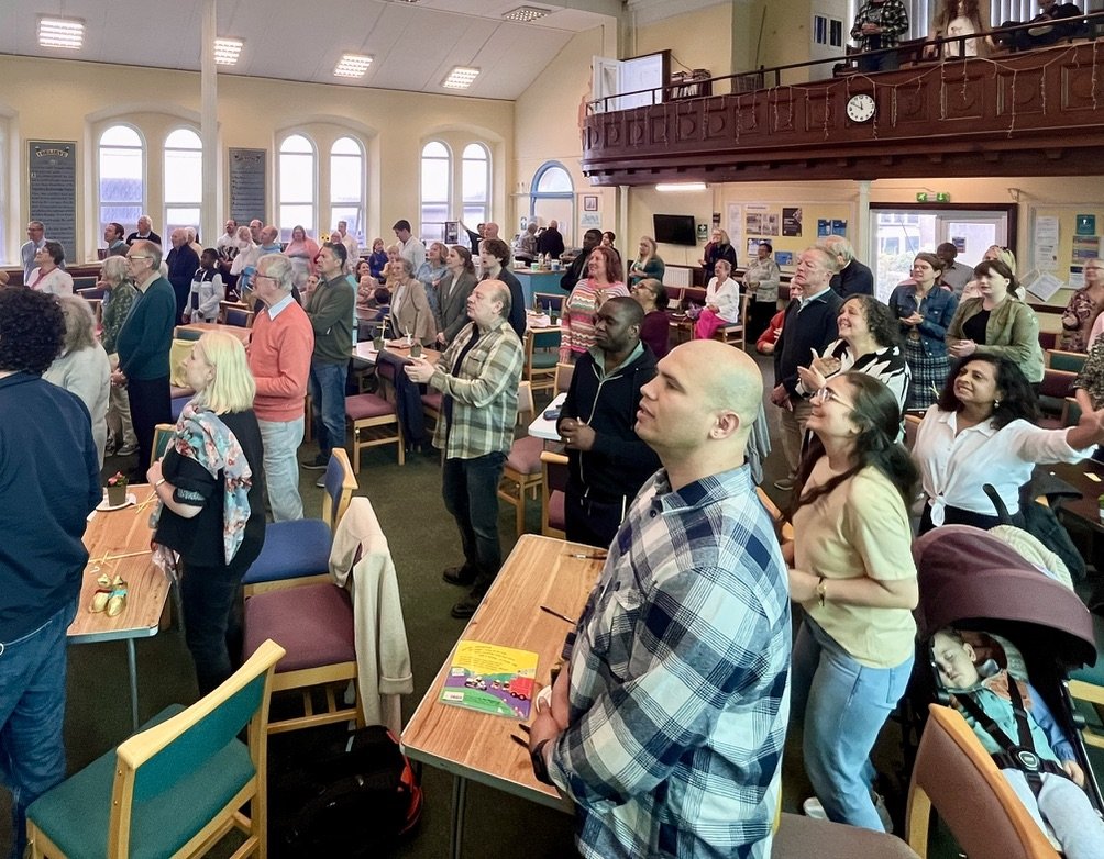 A large group of people standing in a church or meeting hall, some with eyes closed and others with hands raised, engaged in a worship or prayer session.