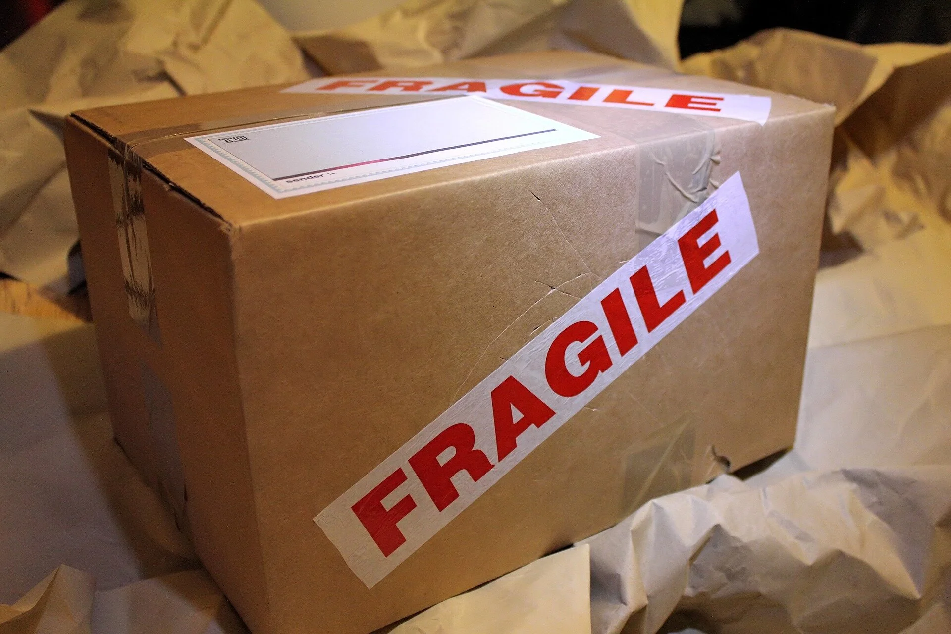 A cardboard box sealed with packing tape and labeled with red and white 'FRAGILE' stickers, surrounded by crumpled packing paper.