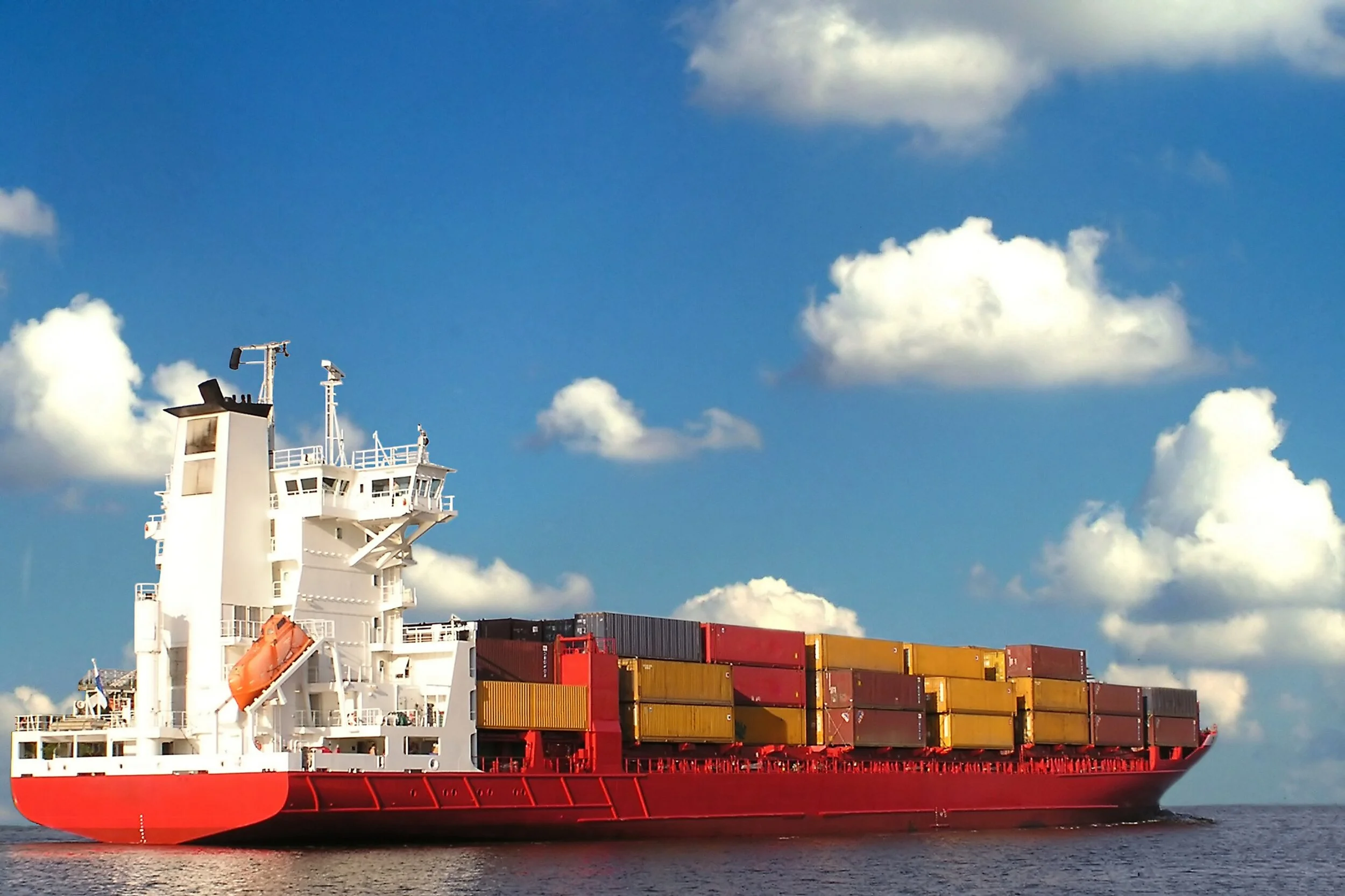 Red cargo ship sailing on open water carrying stacked shipping containers under a blue sky with scattered clouds.
