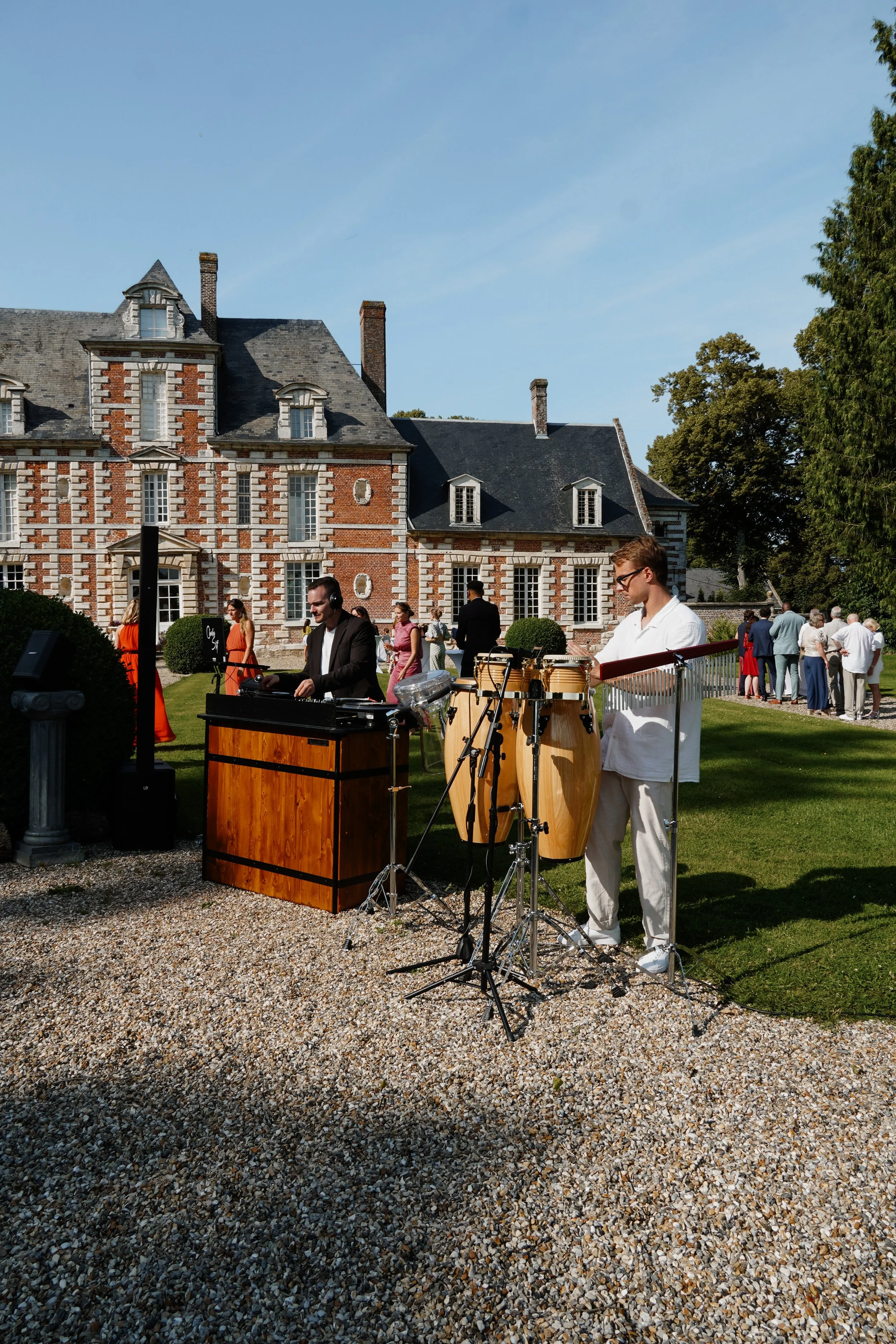 Un groupe de musiciens jouant lors d'un événement en plein air devant un bâtiment en pierre rouge. Certains musiciens portent des écouteurs, d'autres sont en costume ou en vêtements décontractés. Il y a un public en arrière-plan et des arbres en arri