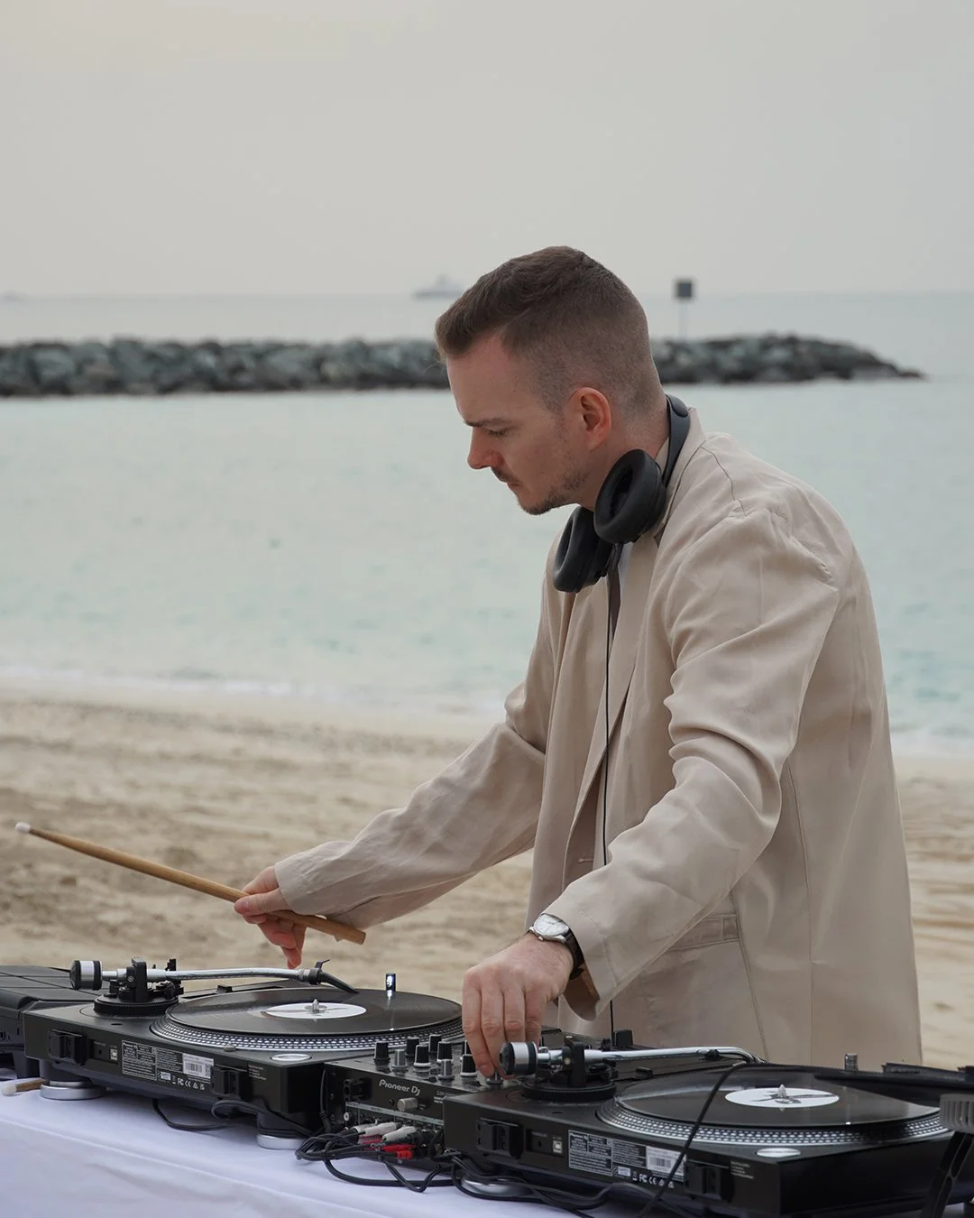 Un DJ joue de la musique sur une plage, portant un casque autour du cou et une veste beige, avec un équipement DJ devant lui, derrière la mer calme et un incident de jetée en pierre.