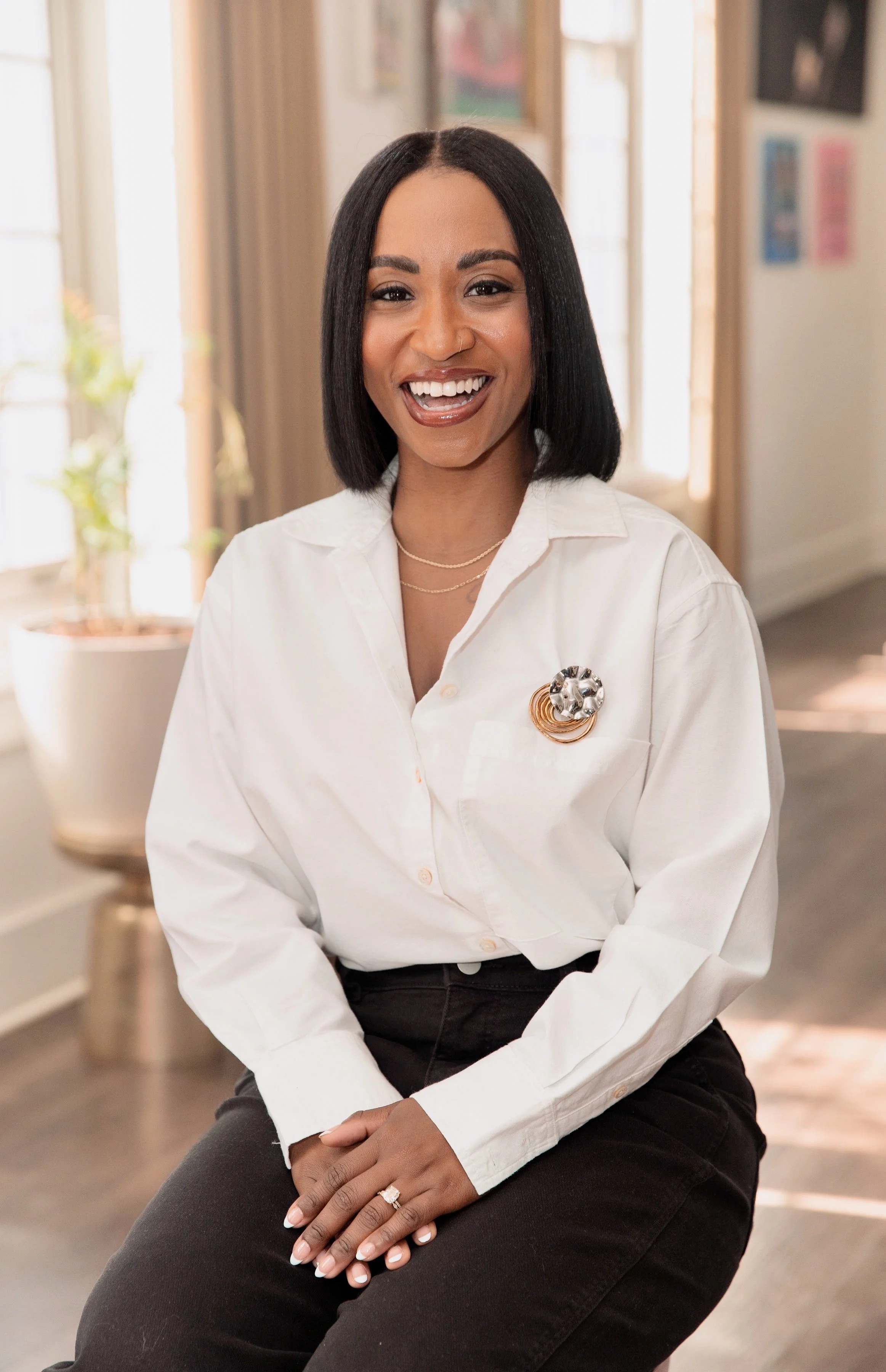 A woman with black hair, wearing a white blouse with a brooch, black pants, and jewelry, sitting indoors and smiling at the camera.