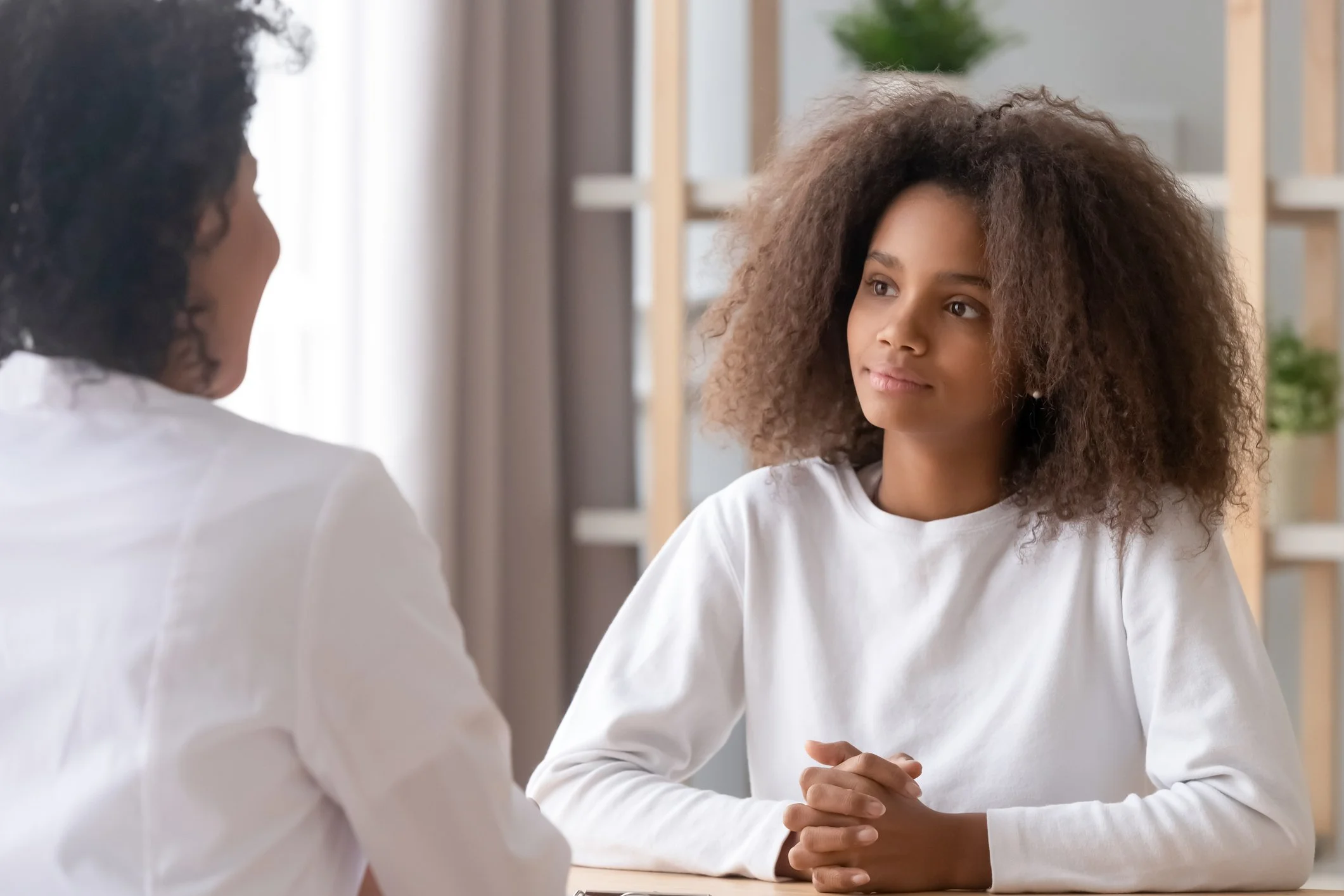 A woman with curly hair listening to a young girl with straight hair in a bright room with bookshelves and plants.
