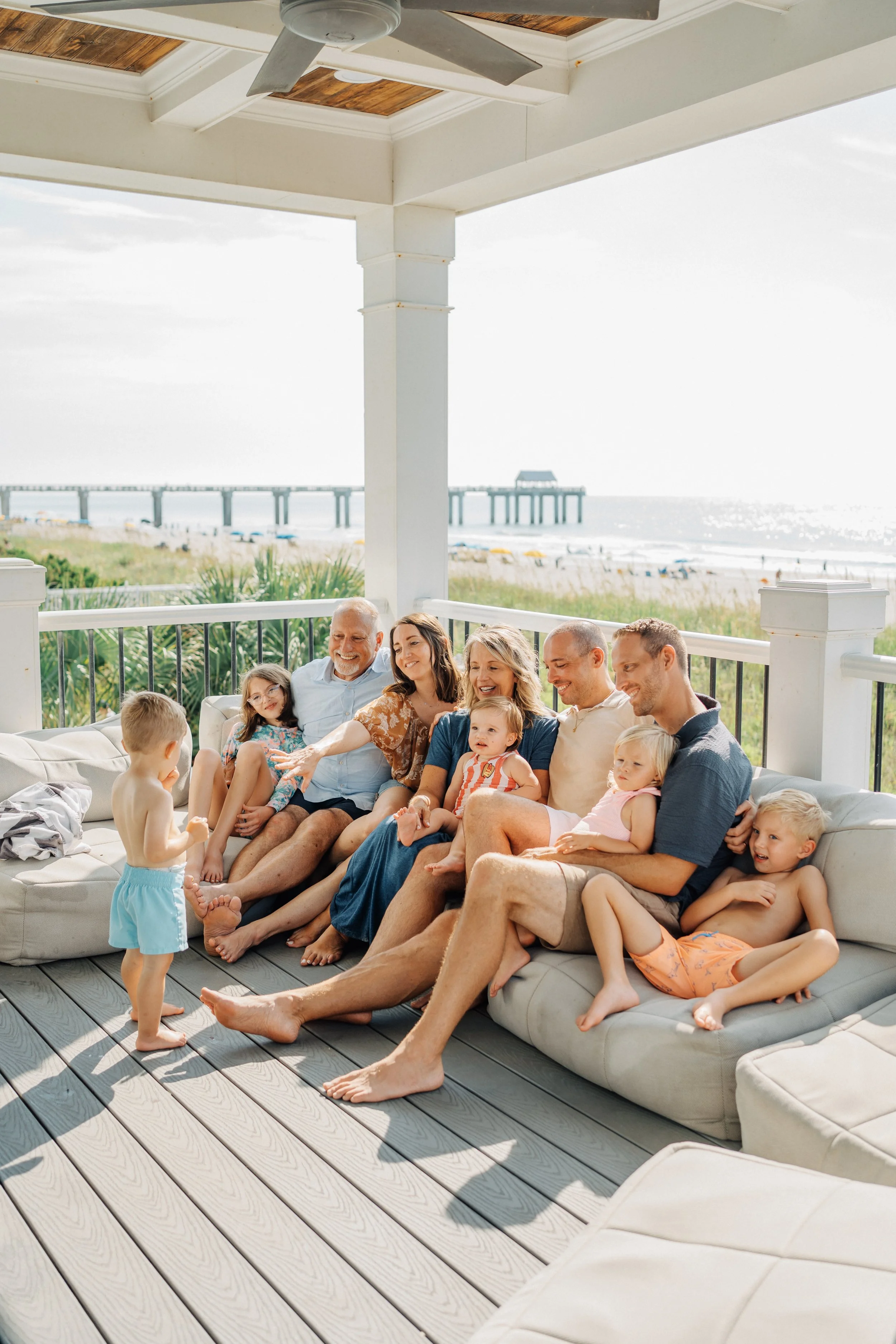 A group of adults and children sitting on a porch sofa by the beach, smiling and interacting, with a pier and ocean in the background on a sunny day.