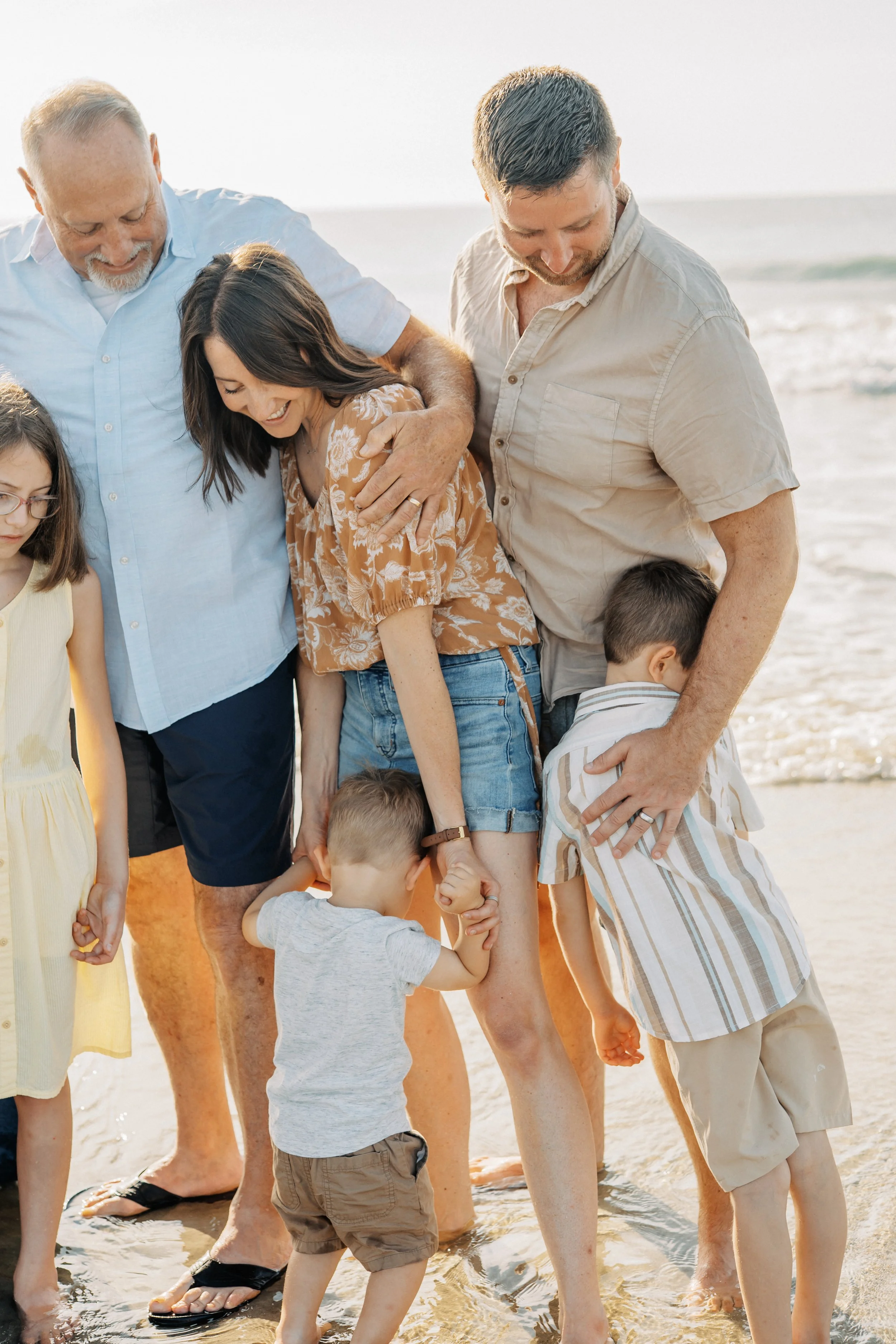 Family reunion on the beach with smiling adults and children, standing in shallow water near the shore, enjoying the sunny weather.