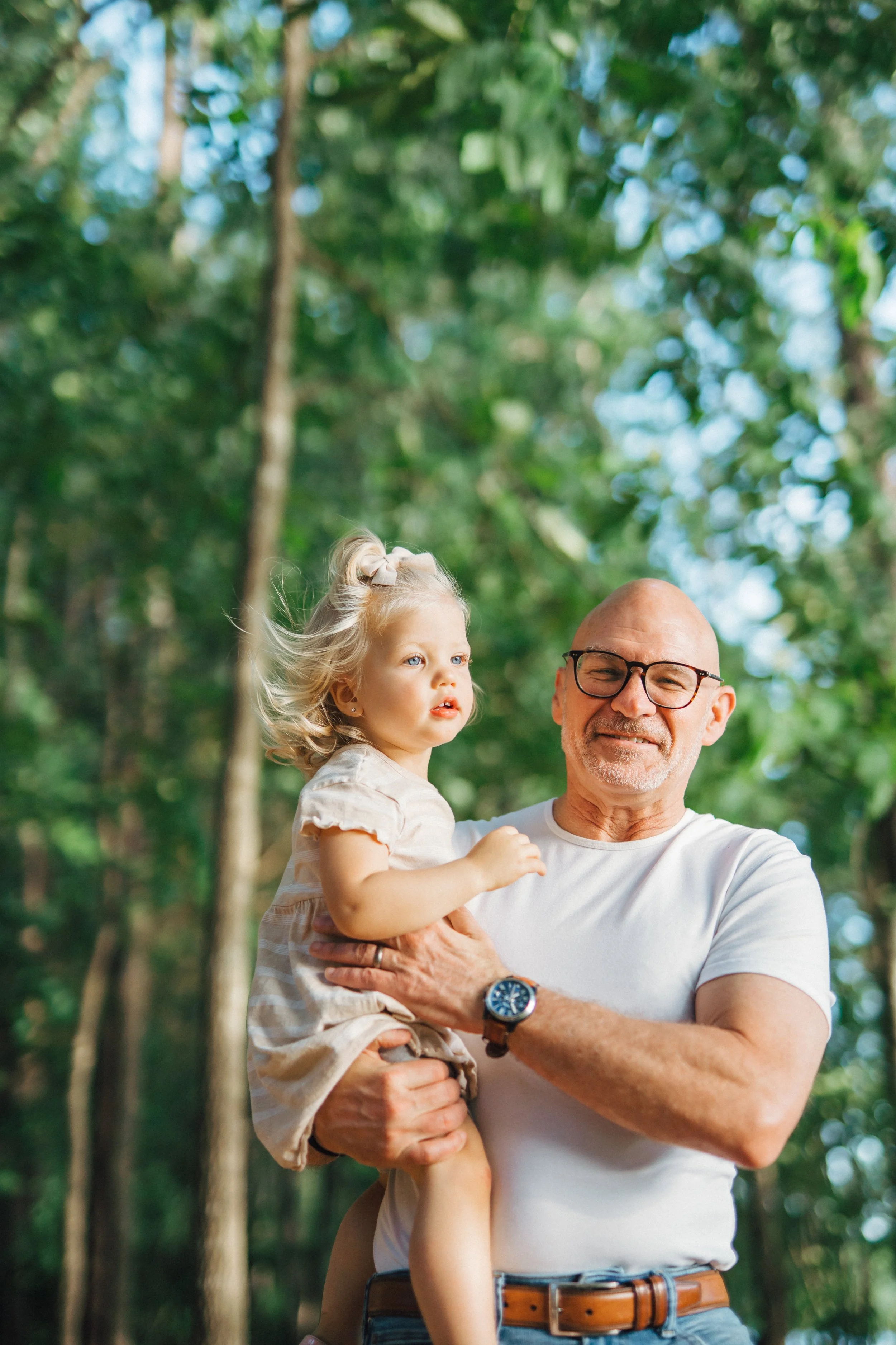 An older man with glasses and a white t-shirt is holding a young girl with blonde hair and a beige dress in a forest with tall trees and green foliage.