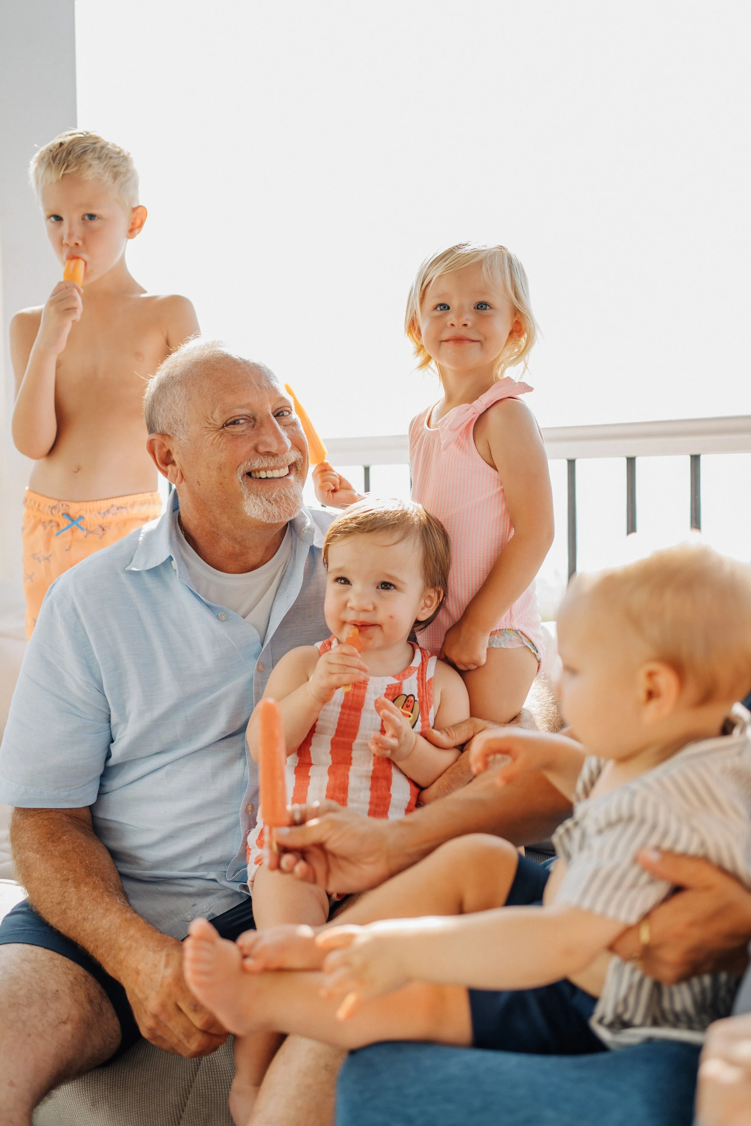 A joyful family of six, including an elderly man surrounded by five young children, with some eating popsicles and others sitting on his lap, in a bright room.