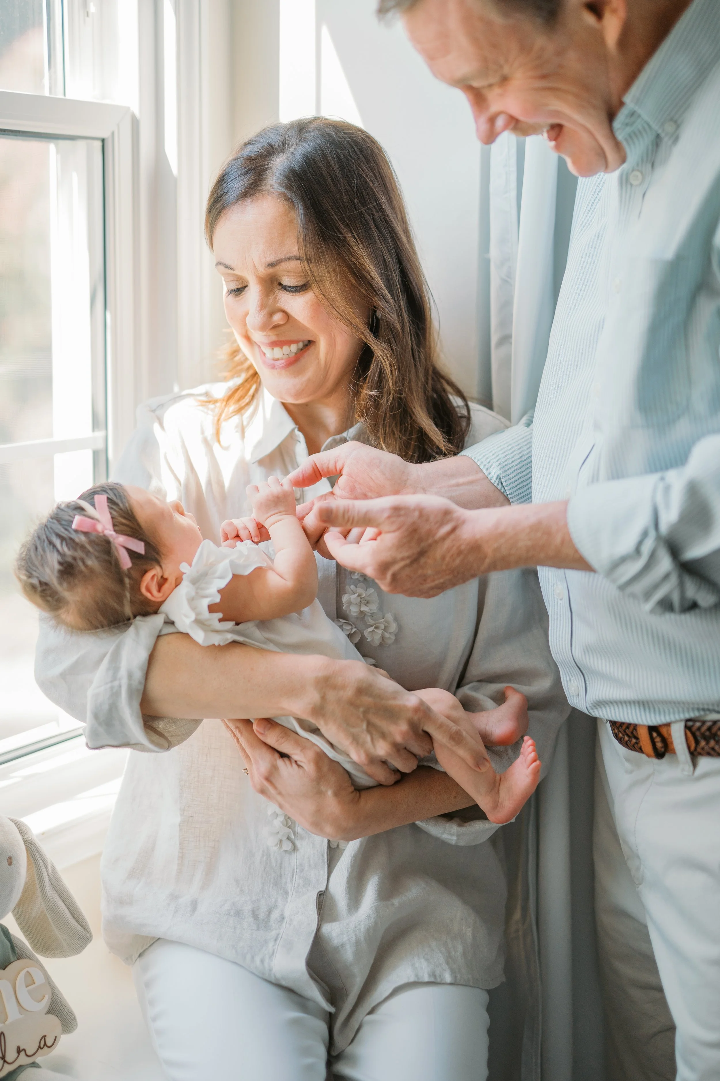 A woman holding a newborn baby girl with a pink bow in her hair, while an elderly man is giving her a small gift or token. They are near a window, smiling and sharing a joyful moment.