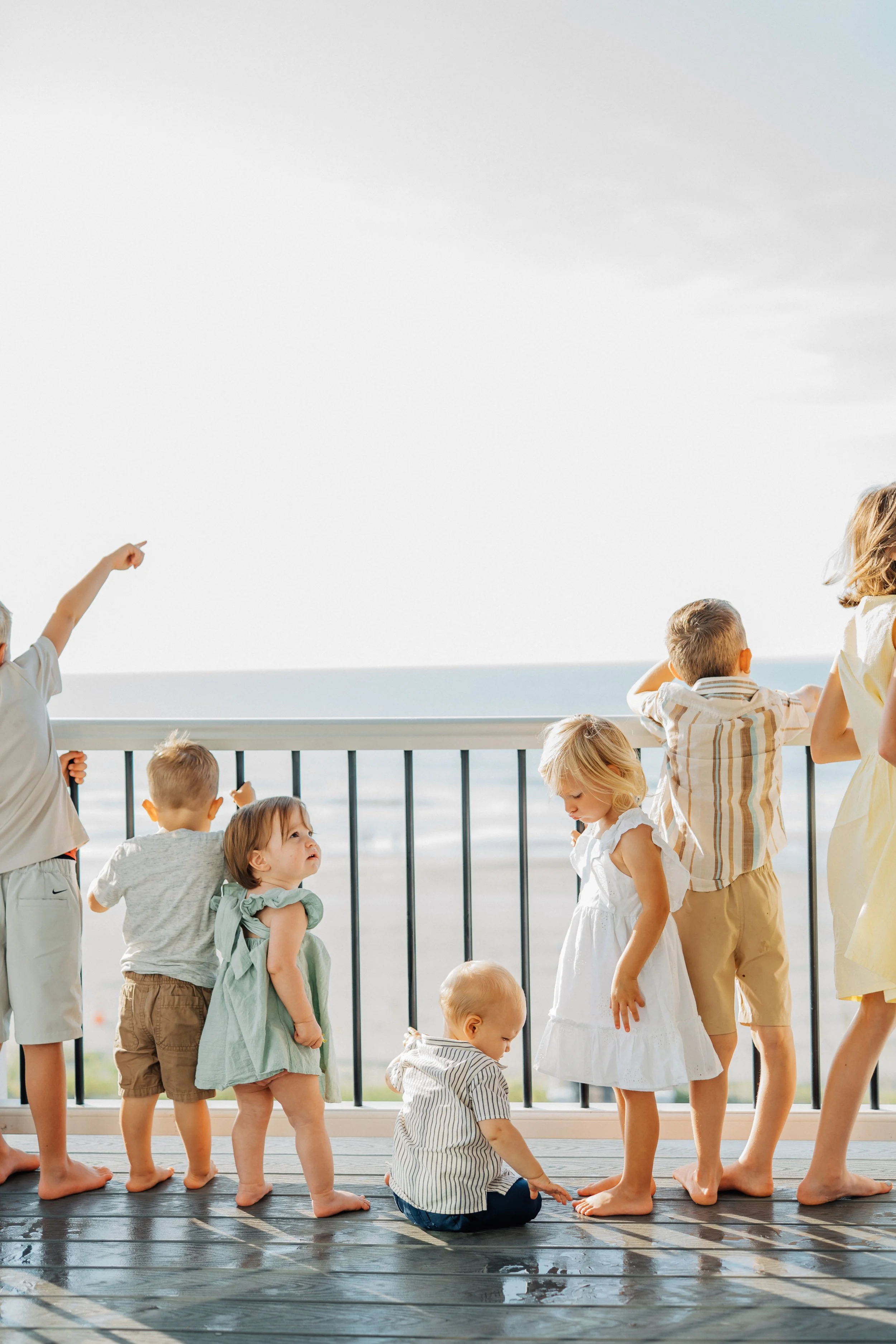 Eight children standing barefoot on a wooden deck overlooking the ocean, some looking out at the sea and others interacting with each other.