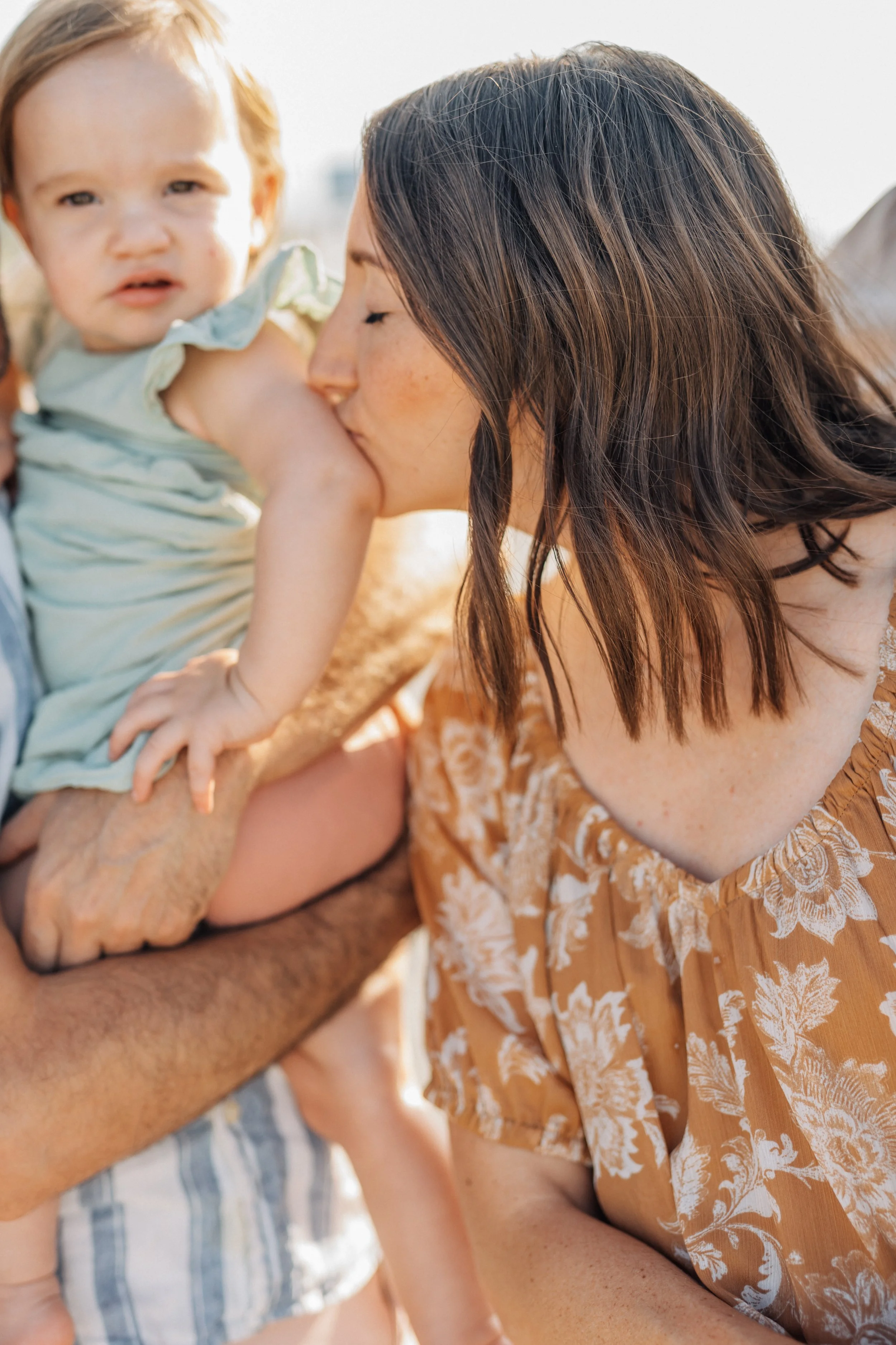 A woman with brown hair kissing a young girl on the cheek, while an older person's arm is holding a baby in the background.