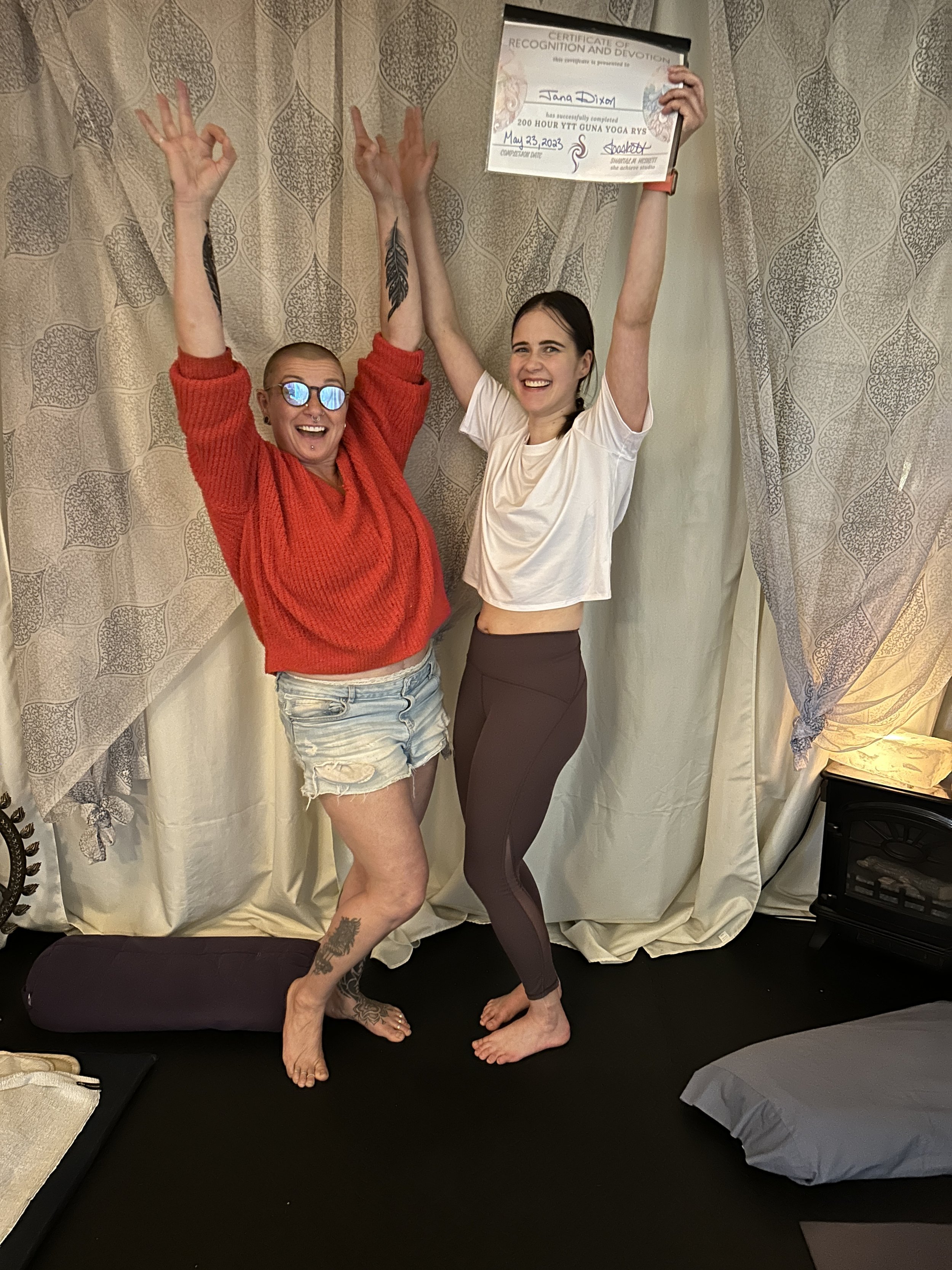 Two women celebrating indoors, one holding a certificate and the other with arms raised. They are smiling and standing on a black mat, with a rolled yoga mat and a pillow visible.