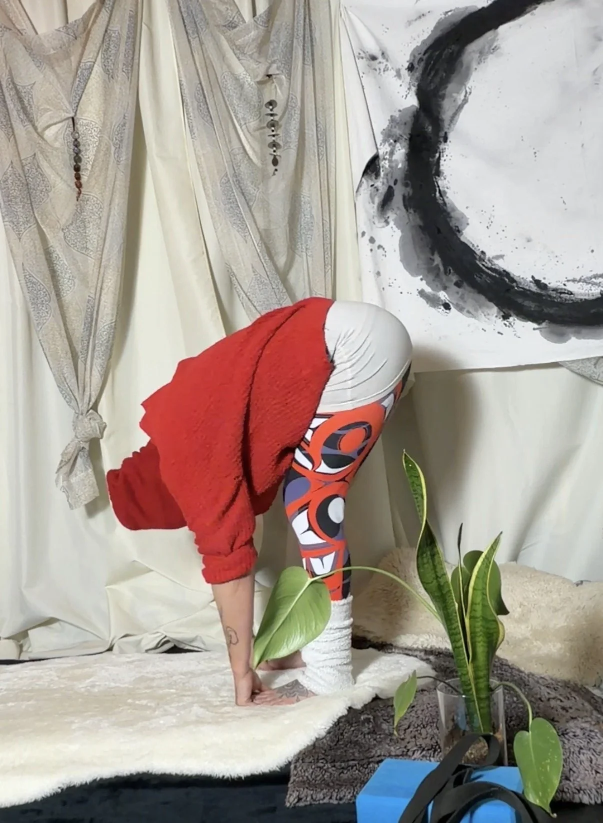 Person in patterned leggings and white socks performing a forward bend yoga pose on a cream-colored rug, with a red hoodie and a large leafy plant nearby, against curtains and wall art background.