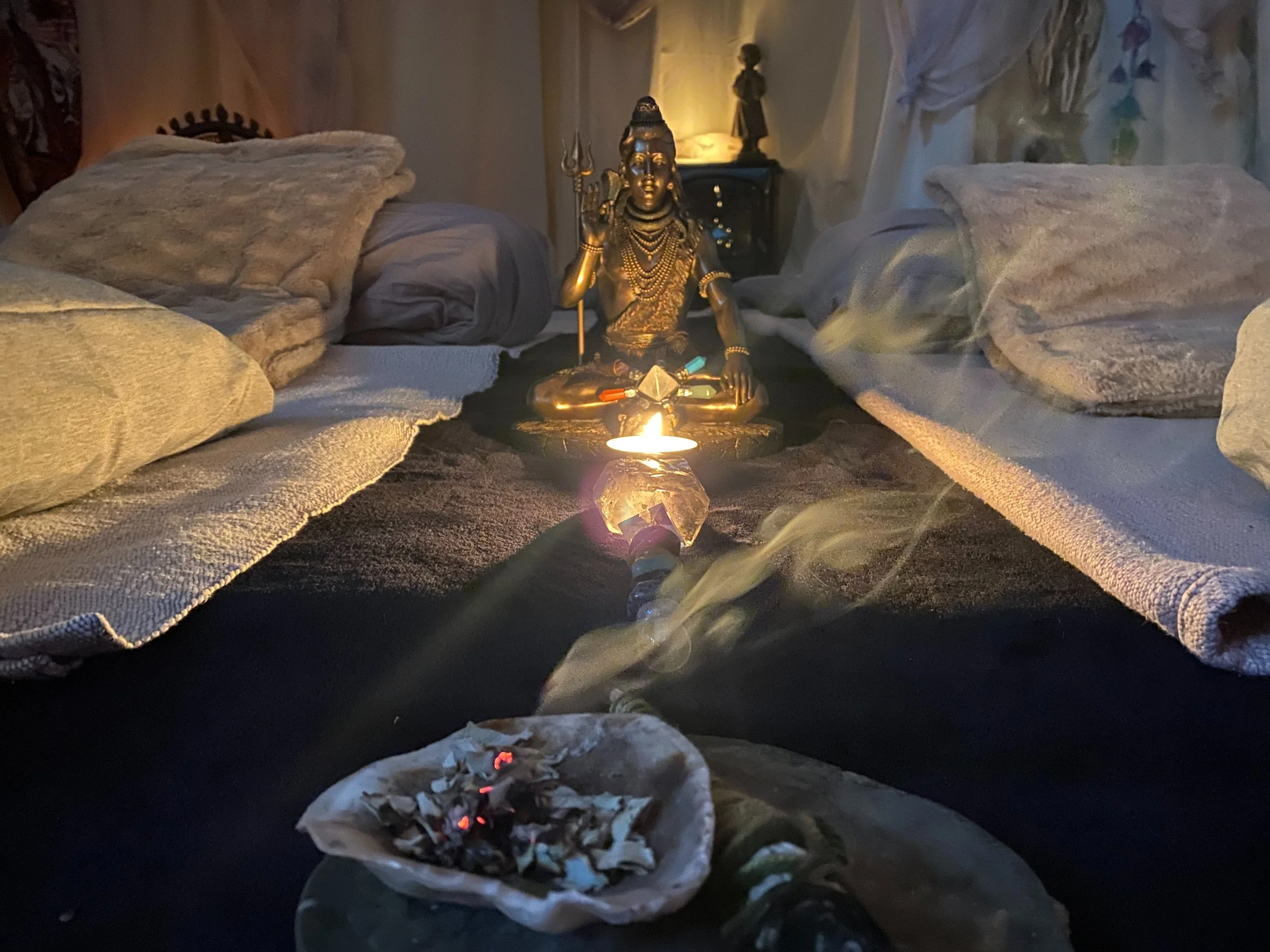 A spiritual or ritual setup with a small lit candle, an incense holder emitting smoke, and a statue of a Hindu deity, possibly Lord Shiva, in the background. The scene has soft lighting with beds or cushions on either side, creating a cozy and sacred atmosphere.