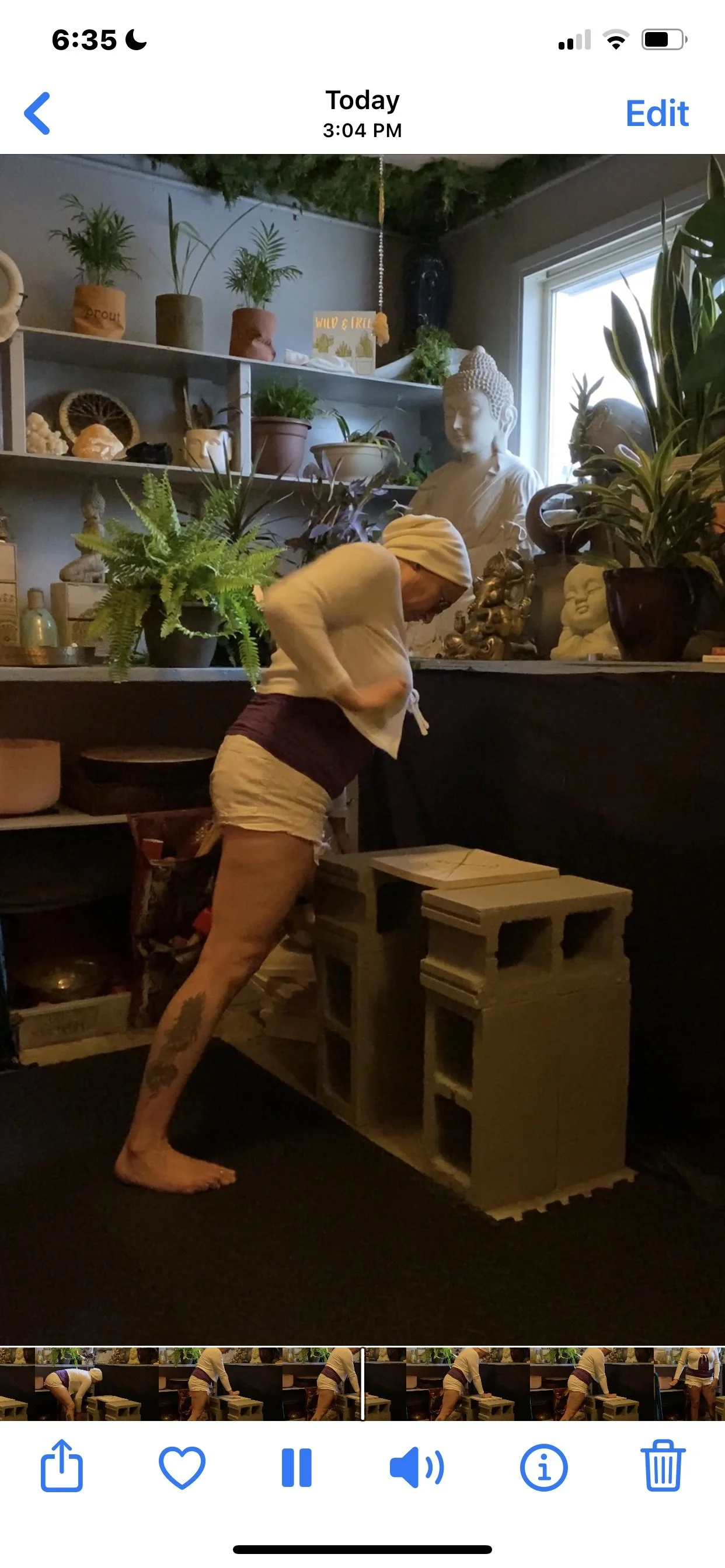 A woman in a white sweater and shorts stands barefoot in a room filled with plants and statues, reaching into a container on a shelf under a window.