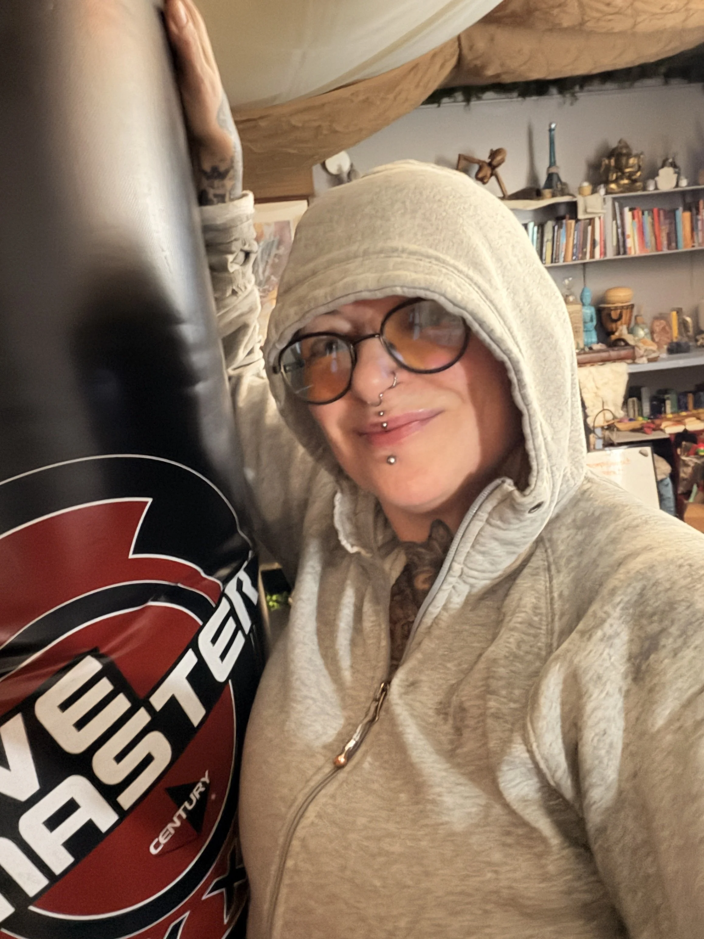 Person wearing glasses, a grey hoodie, and multiple facial piercings, taking a selfie with an object that has a logo reading 'V8 VESTA CENTURY'.