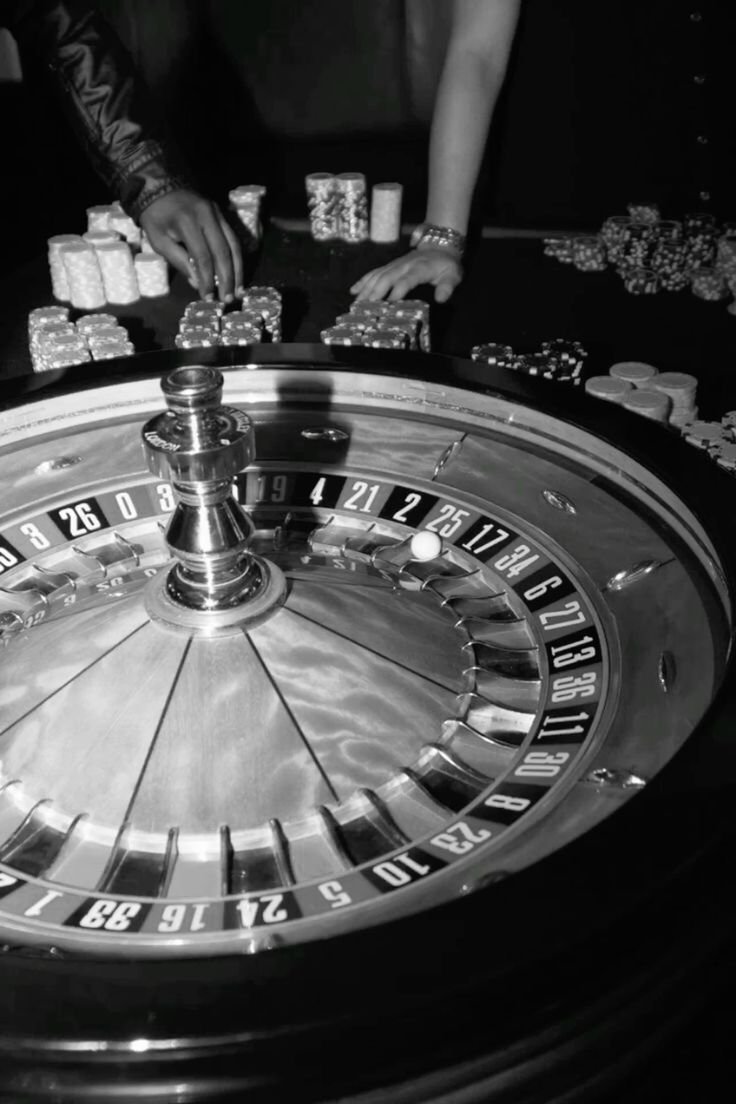 Black and white photo of a roulette wheel in a casino with players placing chips on the table.