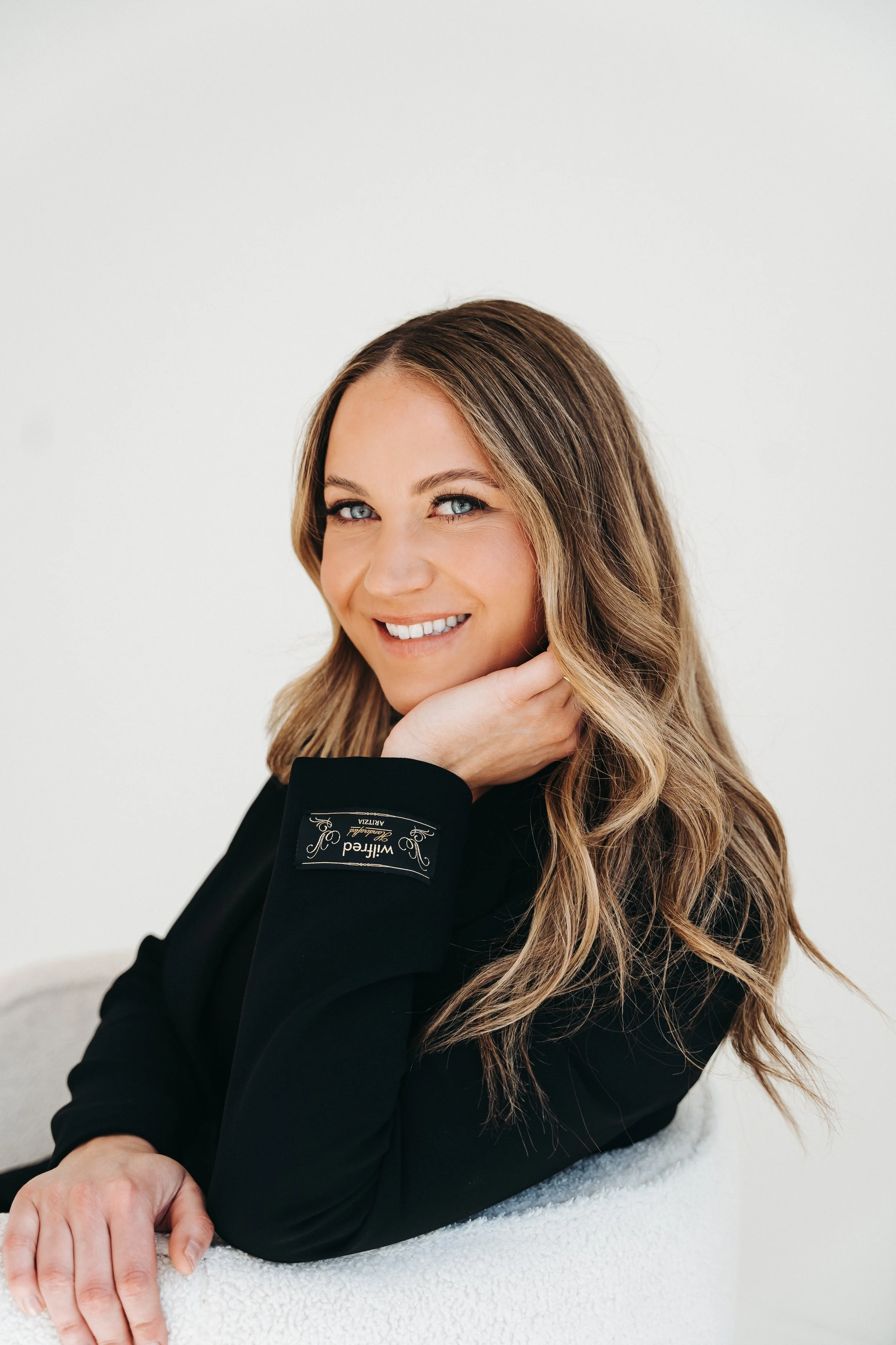 A woman with long, wavy blonde hair and blue eyes, smiling and resting her chin on her hand, wearing a black blazer with a label reading "Wilfred" on the sleeve, sitting on a white surface against a plain white background.